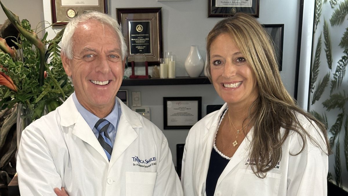 A portrait of Dr. Frederick Solomon, D.M.D., and Danielle Solomon, R.D.H., smiling in a dental office setting with awards displayed in the background.