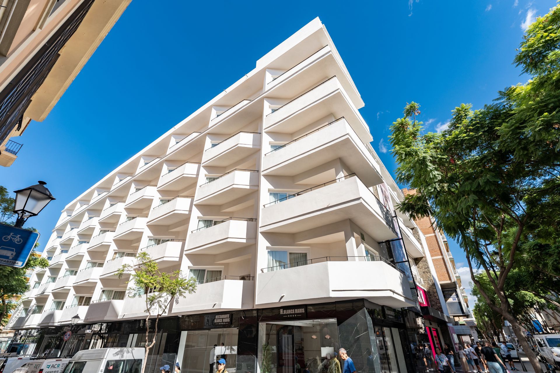 Edificio blanco con balcones, tiendas abajo, cielo azul.