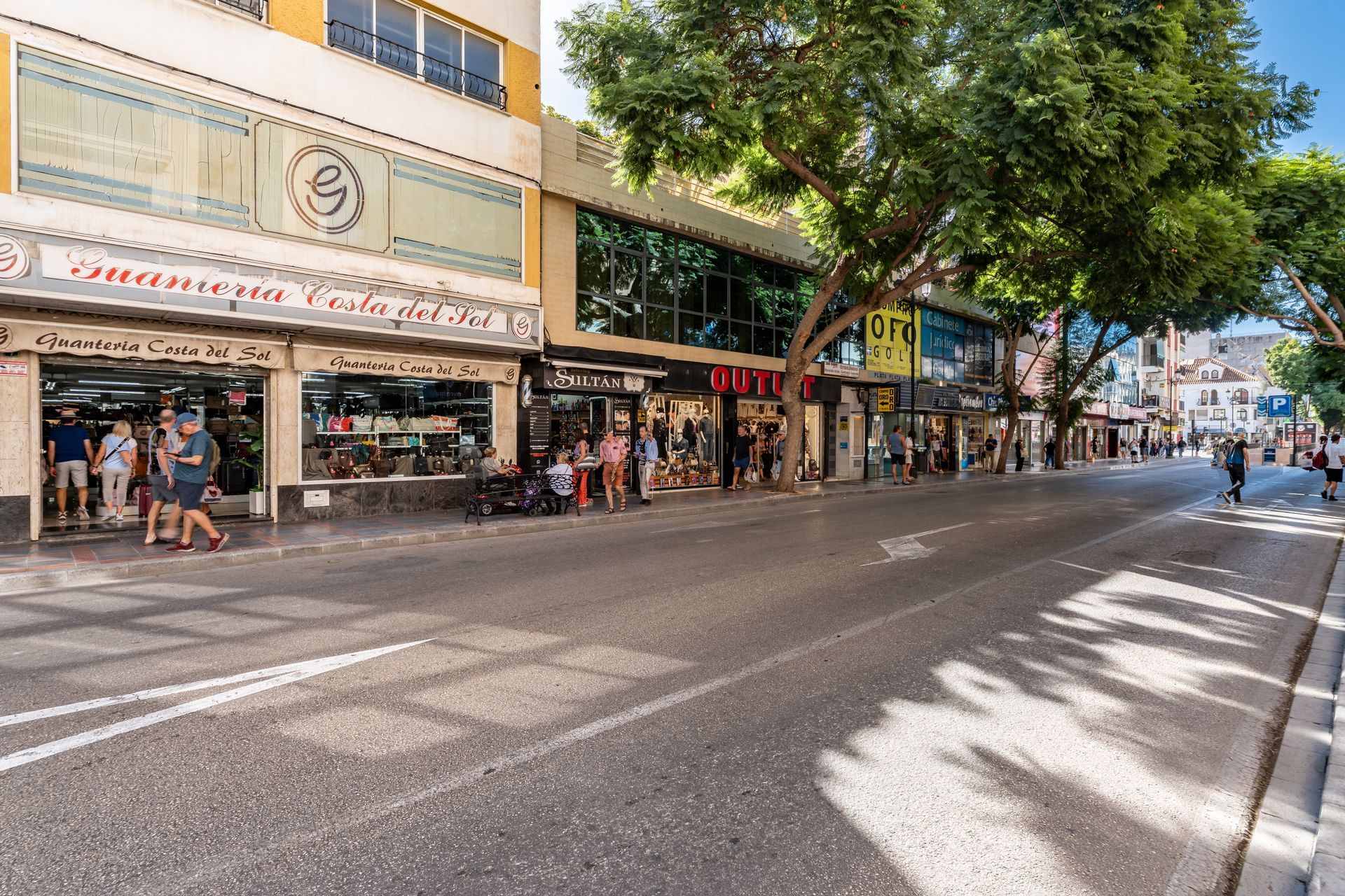 Calle con tiendas, gente caminando. Día soleado, edificios a ambos lados, árboles.