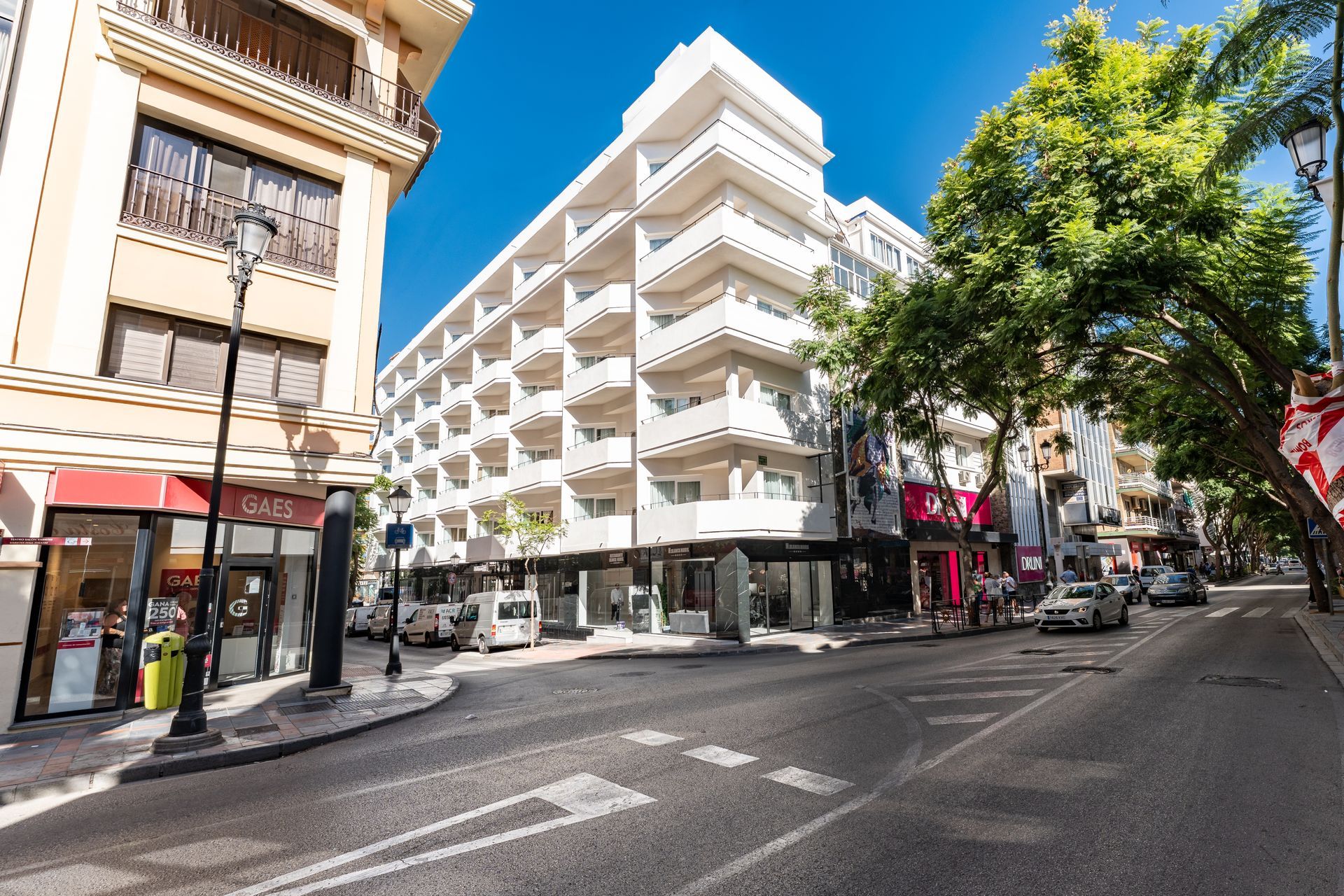 Vista de la calle de un edificio blanco con balcones, tiendas, coches y árboles en un día soleado.