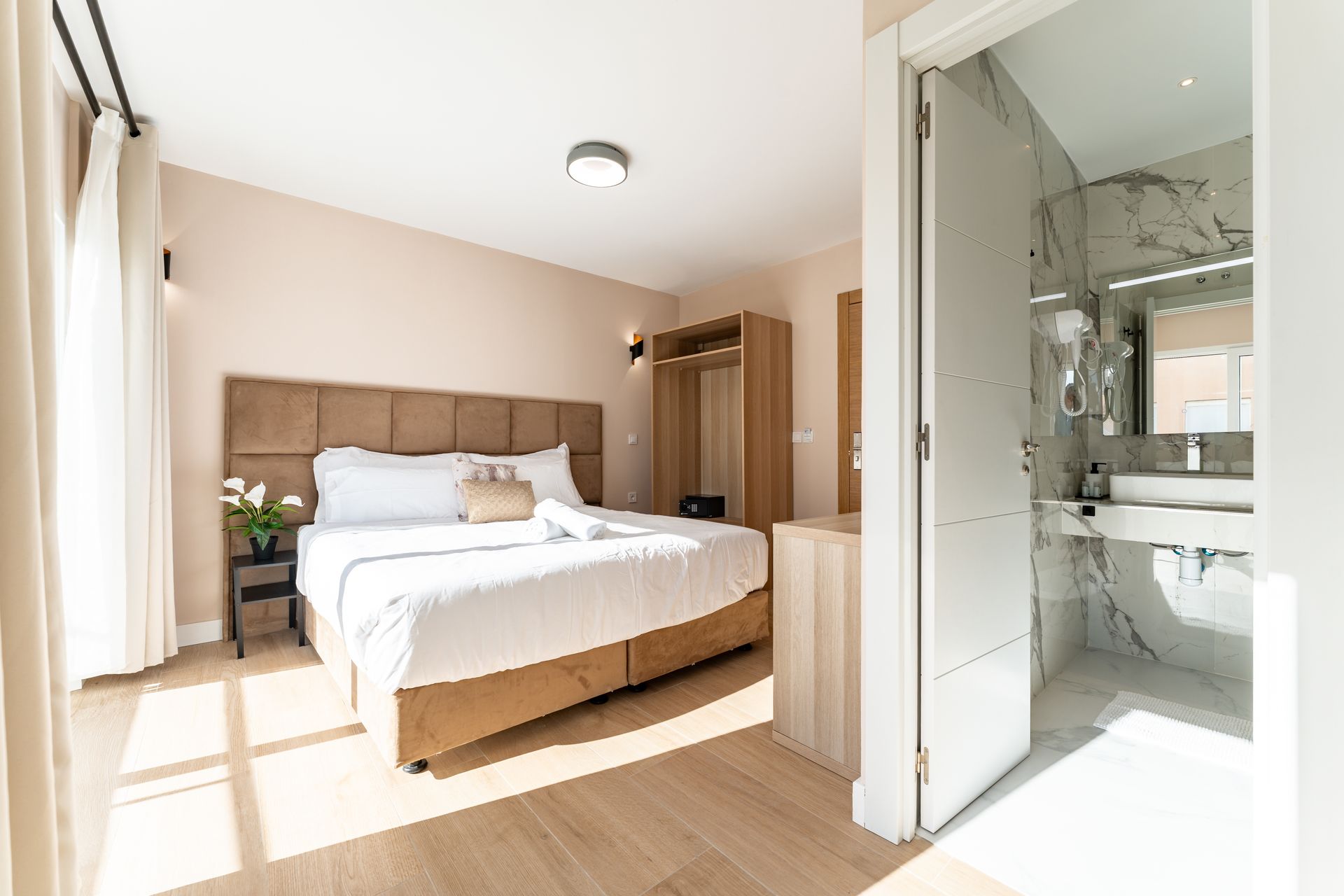 Dormitorio con cama, armario y baño a la vista. Paredes y suelo de color claro, con abundante luz natural.