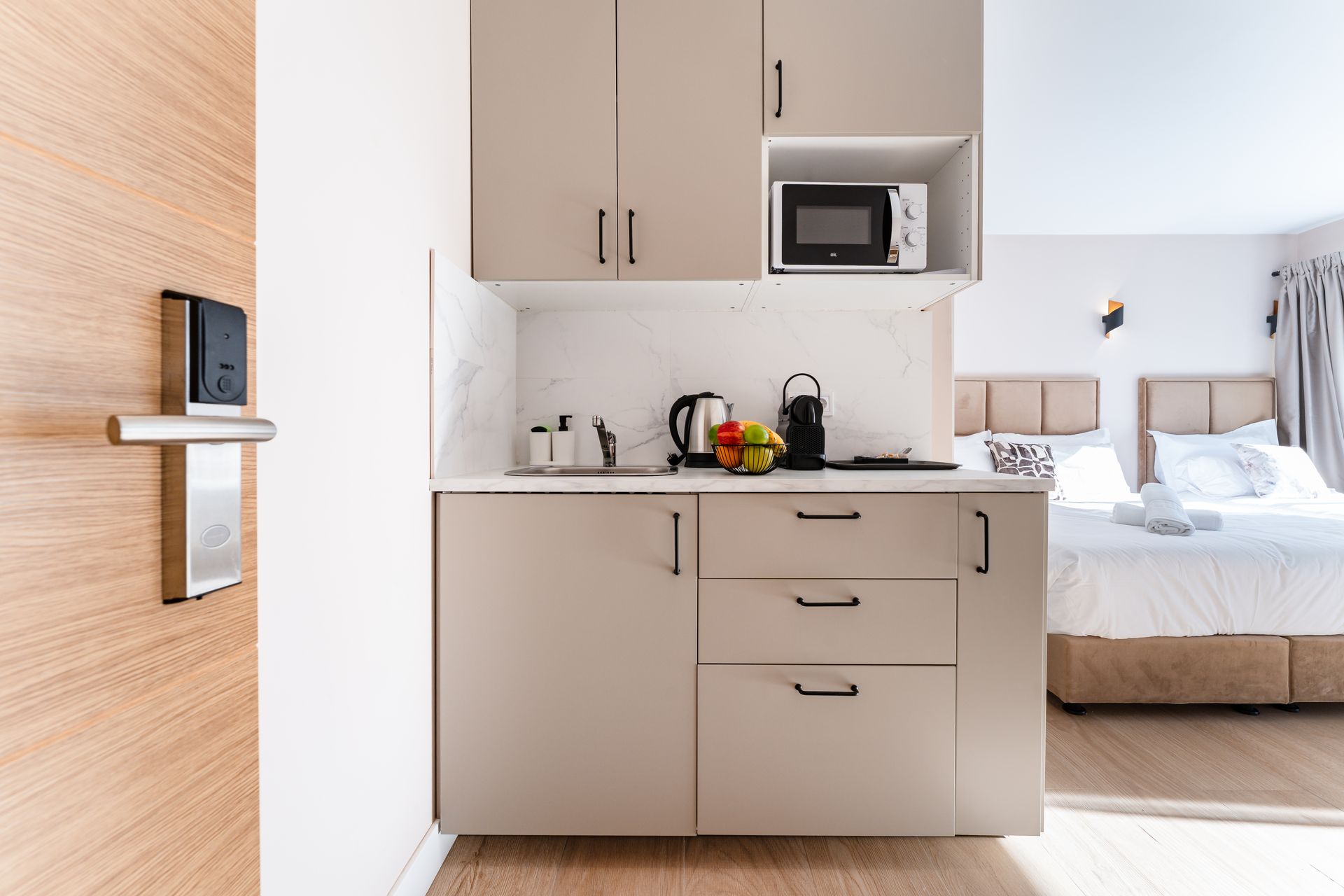 Pequeña cocina en una habitación de hotel con microondas, cafetera y fruta, junto a la cama.