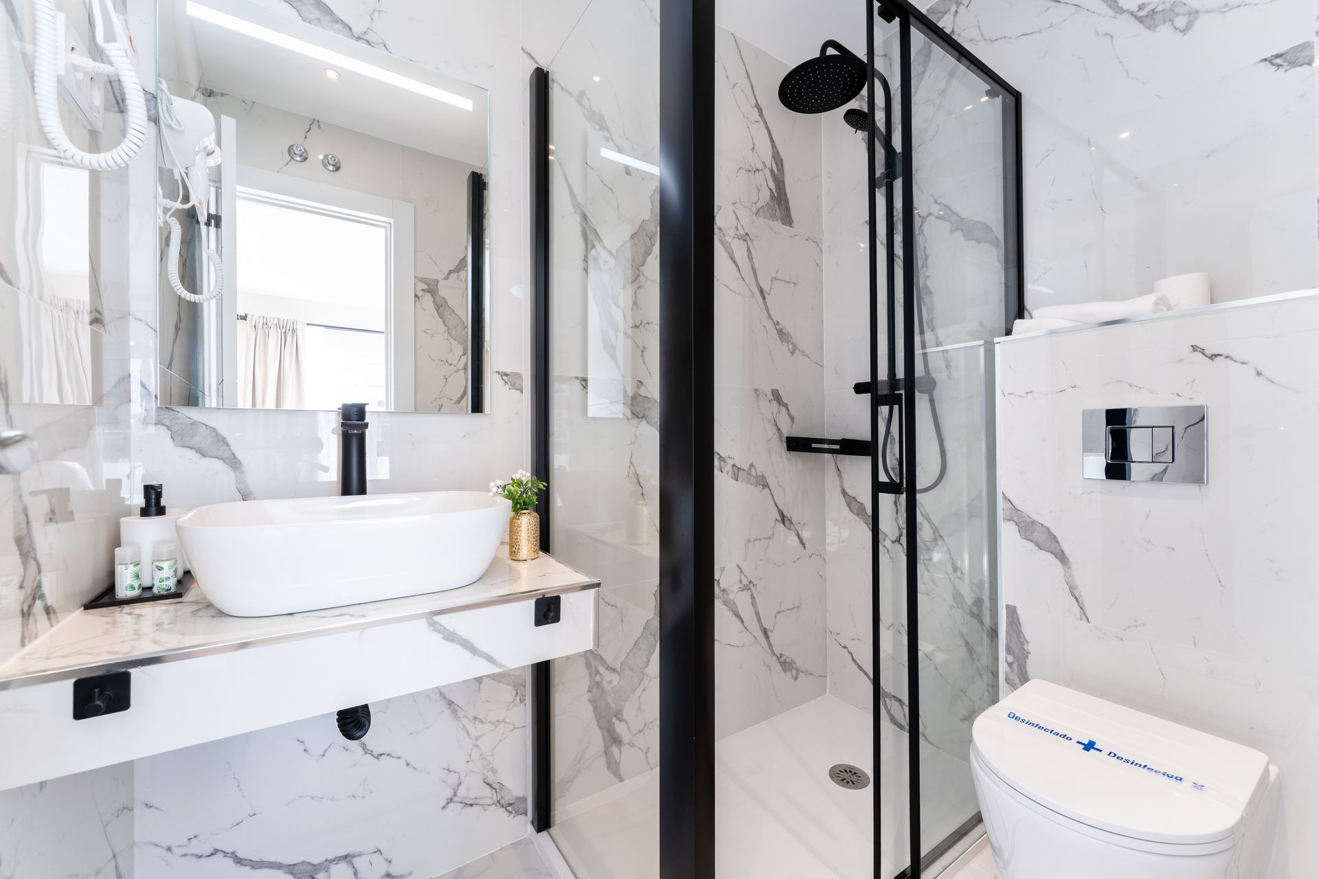 Baño moderno con paredes de mármol, lavabo sobre encimera y ducha con marco negro.