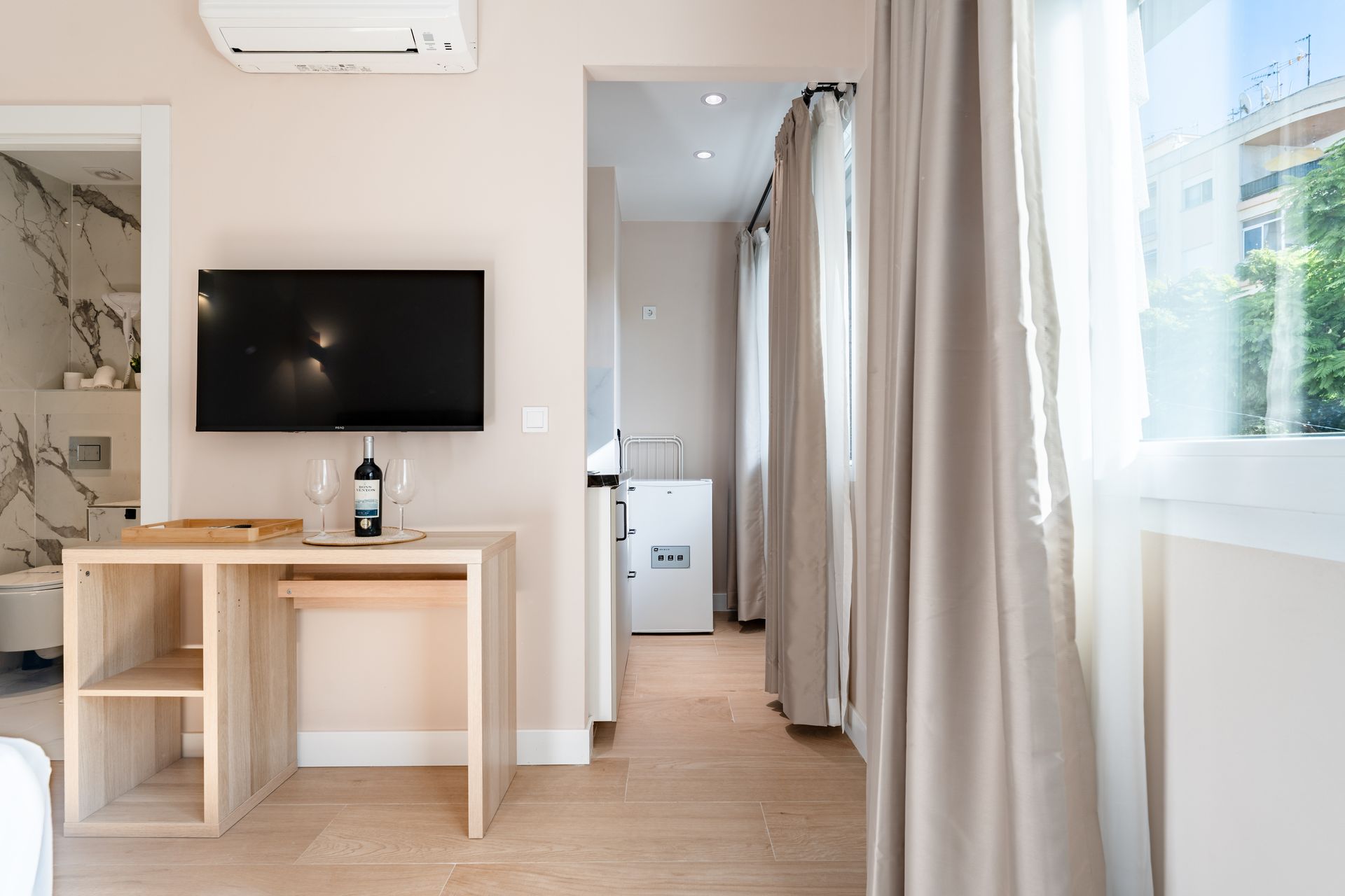 Una habitación luminosa con un escritorio, un televisor y una puerta abierta a otra habitación.