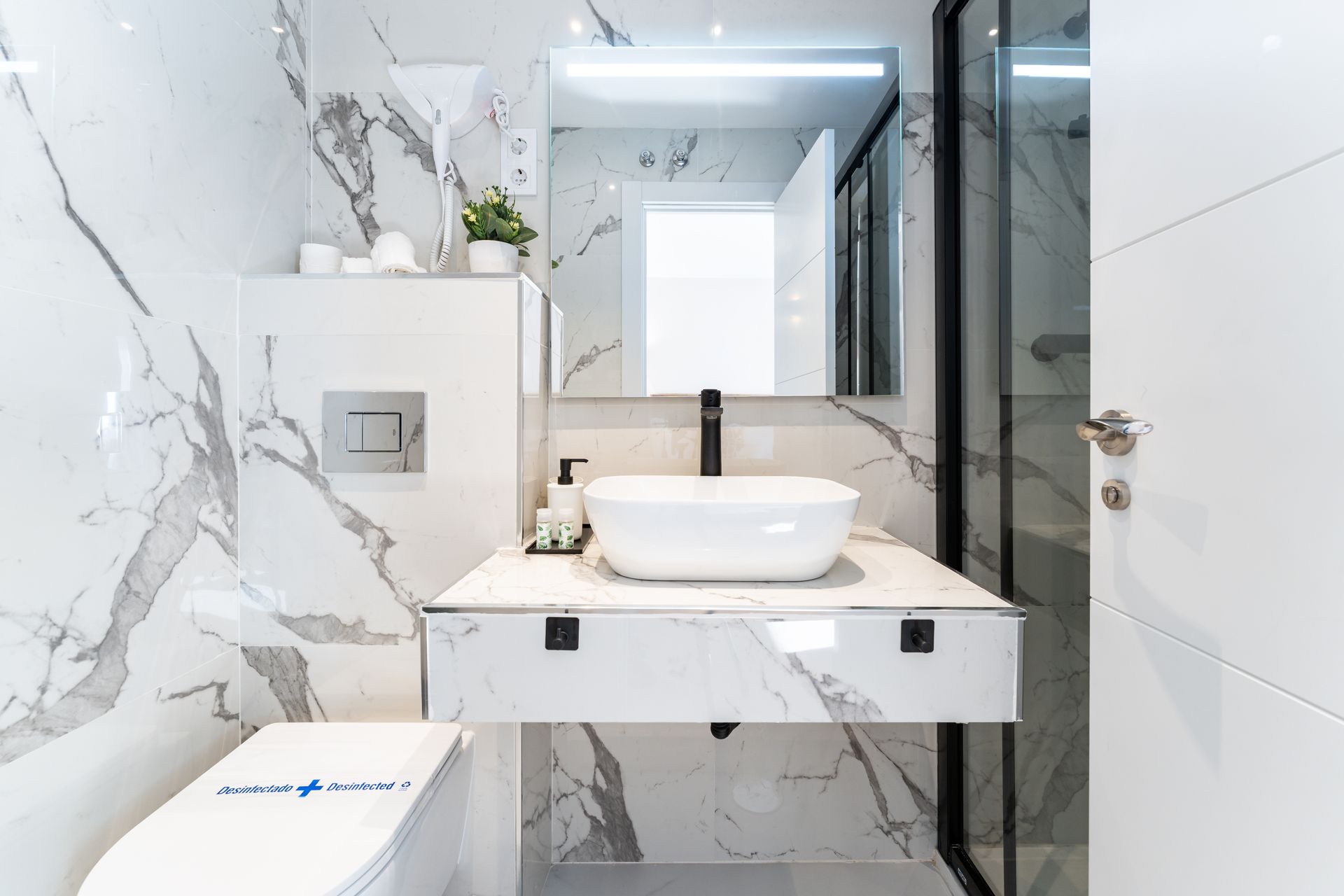 Baño moderno blanco con azulejos de mármol, tocador suspendido, lavabo sobre encimera, grifo negro y ducha.