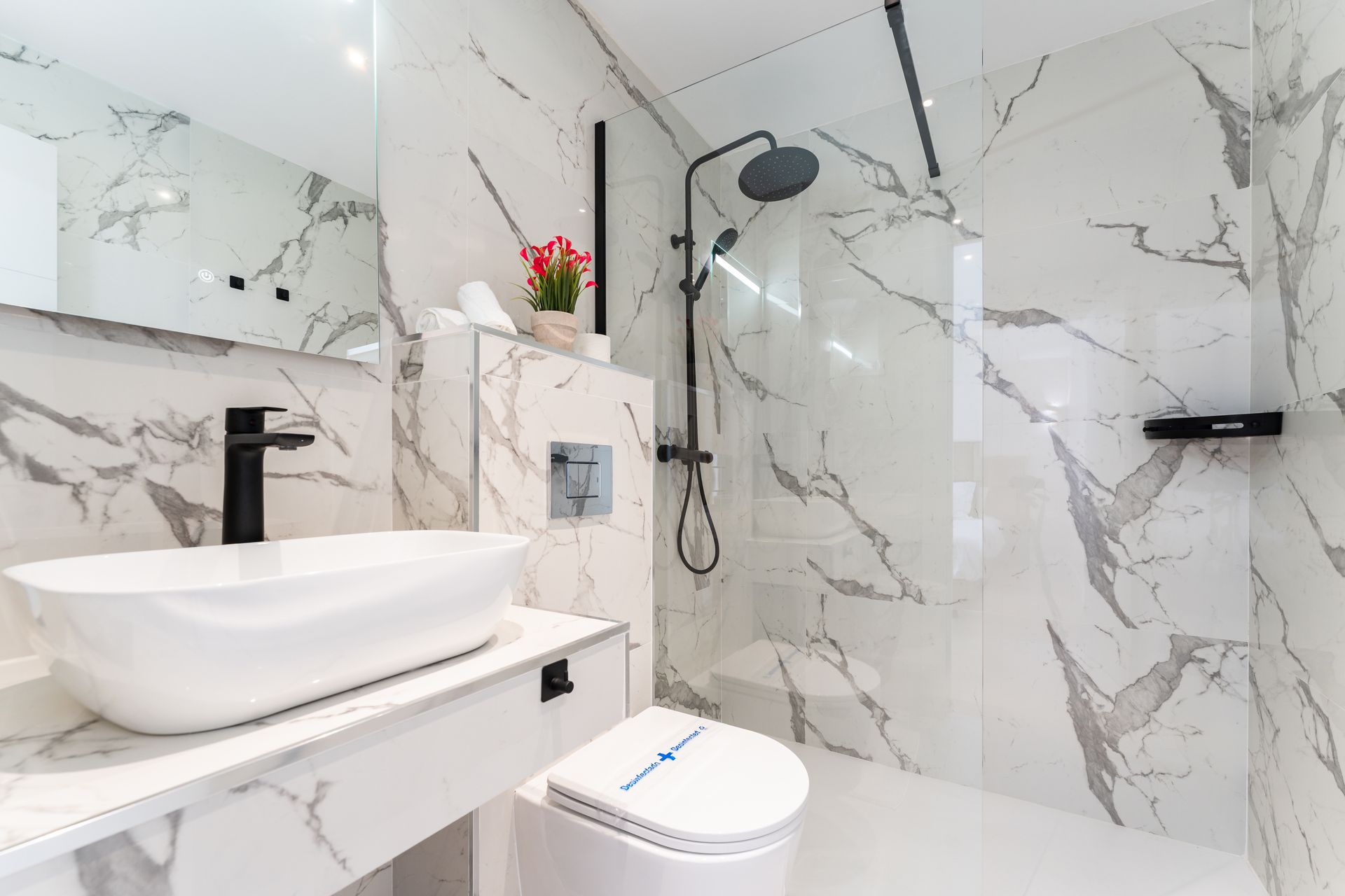 Baño moderno con paredes efecto mármol, lavabo blanco y grifería negra. Incluye ducha, inodoro y espejo.