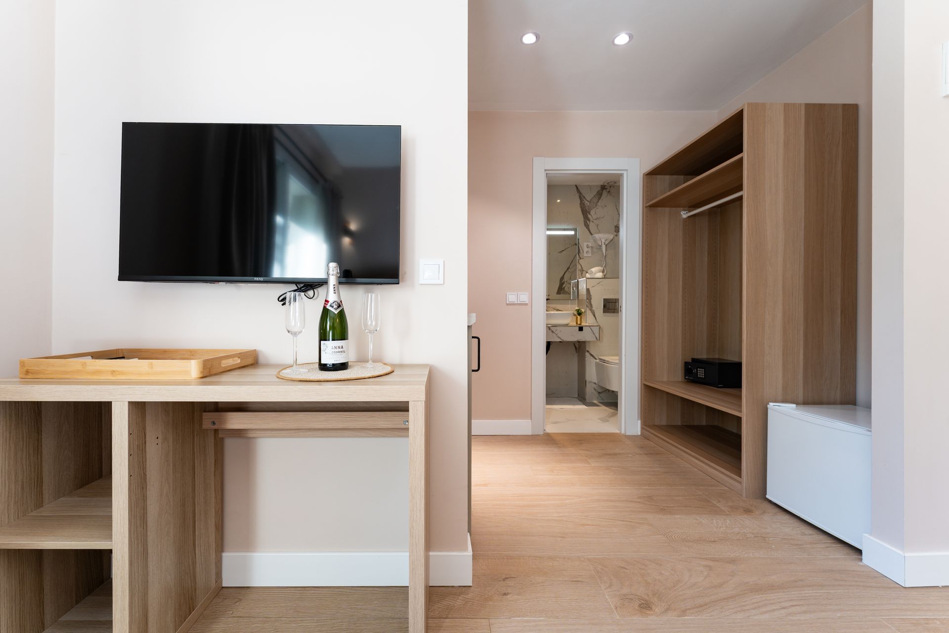 Interior de una habitación de hotel en tonos claros: un televisor sobre un escritorio con champán, un armario.