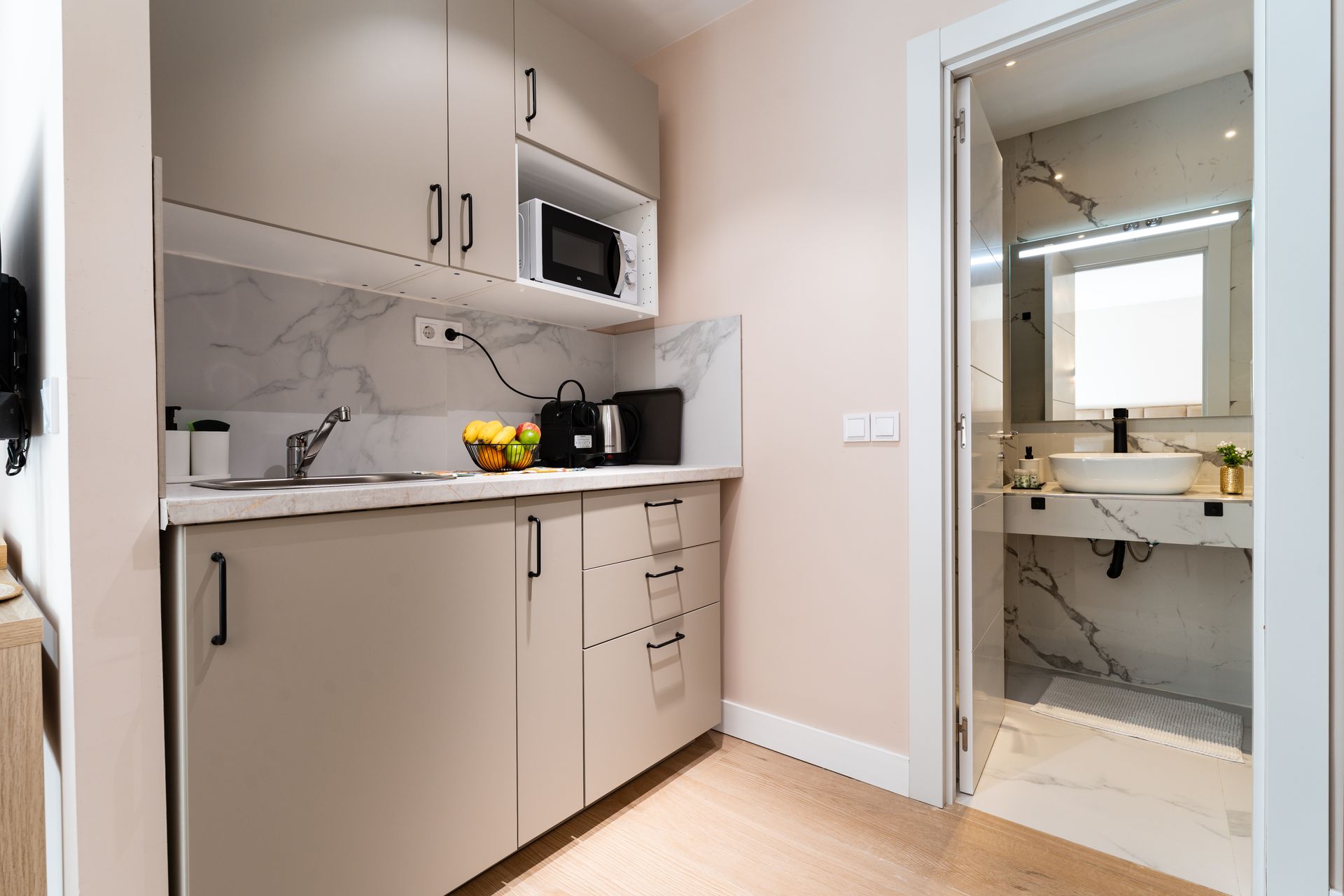 Pequeña cocina con armarios color beige, microondas y fregadero, conectada a un baño con lavabo y espejo.