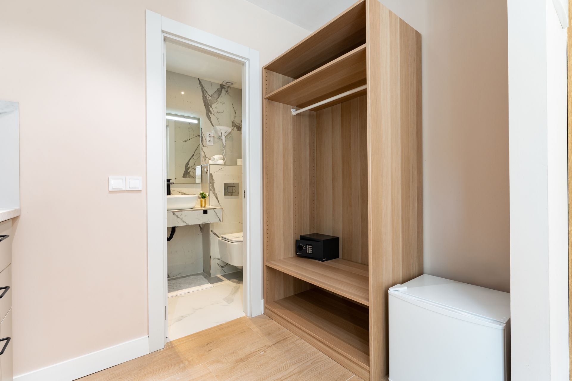 Armario de madera abierto junto a la puerta del baño. Paredes color beige, suelo de madera clara. Mininevera.