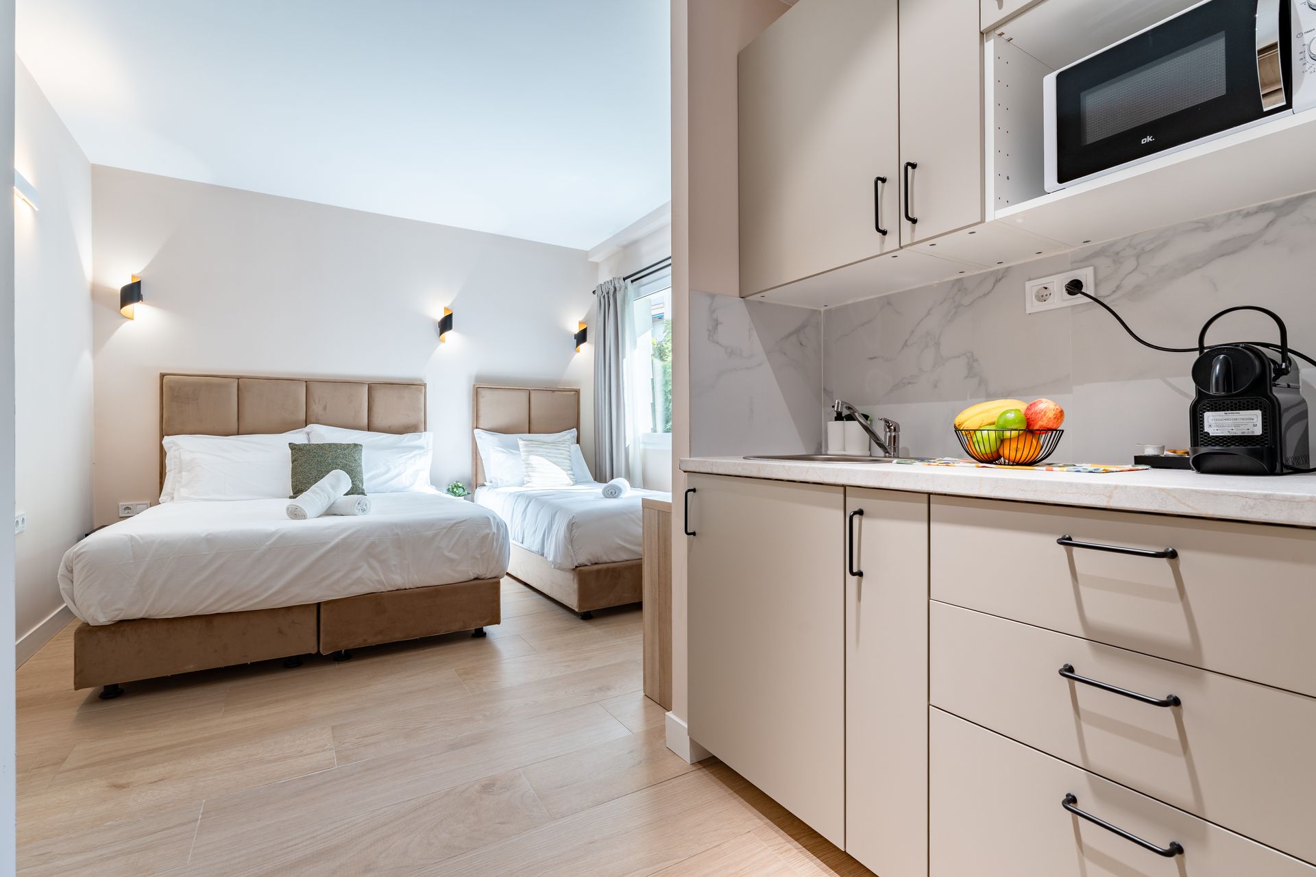 Dormitorio y cocina americana en tonos neutros. Dos camas, pequeña cocina con microondas, fregadero y frutero.