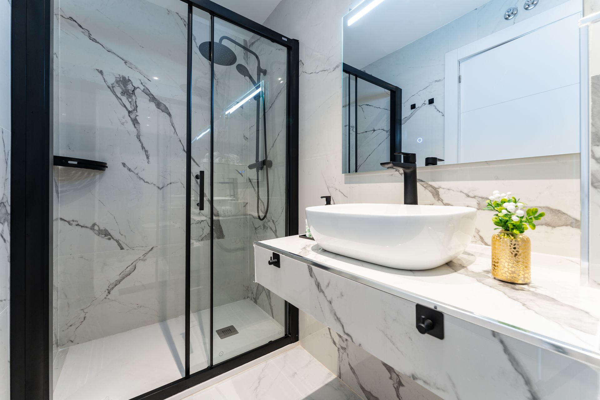 Baño moderno con paredes de mármol, ducha con marco negro, lavabo sobre encimera blanco y detalles dorados.