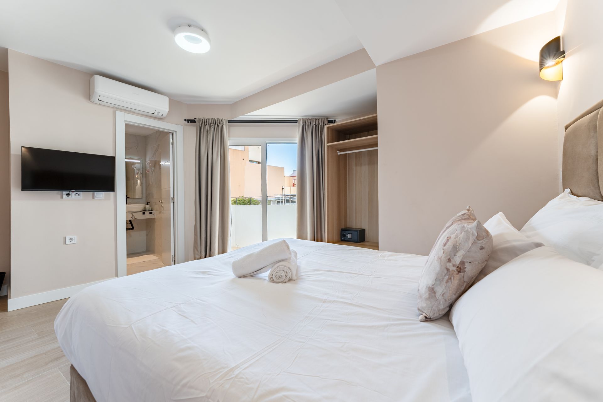 Dormitorio con una gran cama blanca, TV, aire acondicionado y vistas a un balcón.