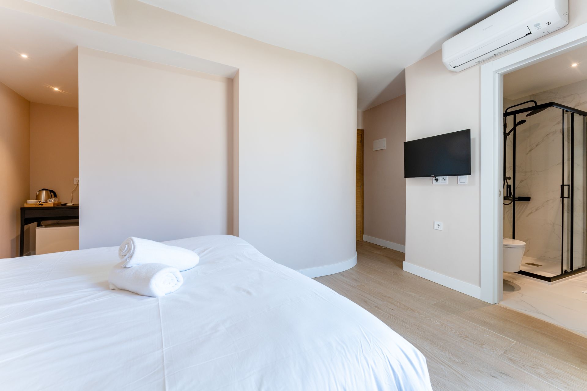 Dormitorio con ropa de cama blanca, televisor en la pared, baño con mampara de ducha negra visible.