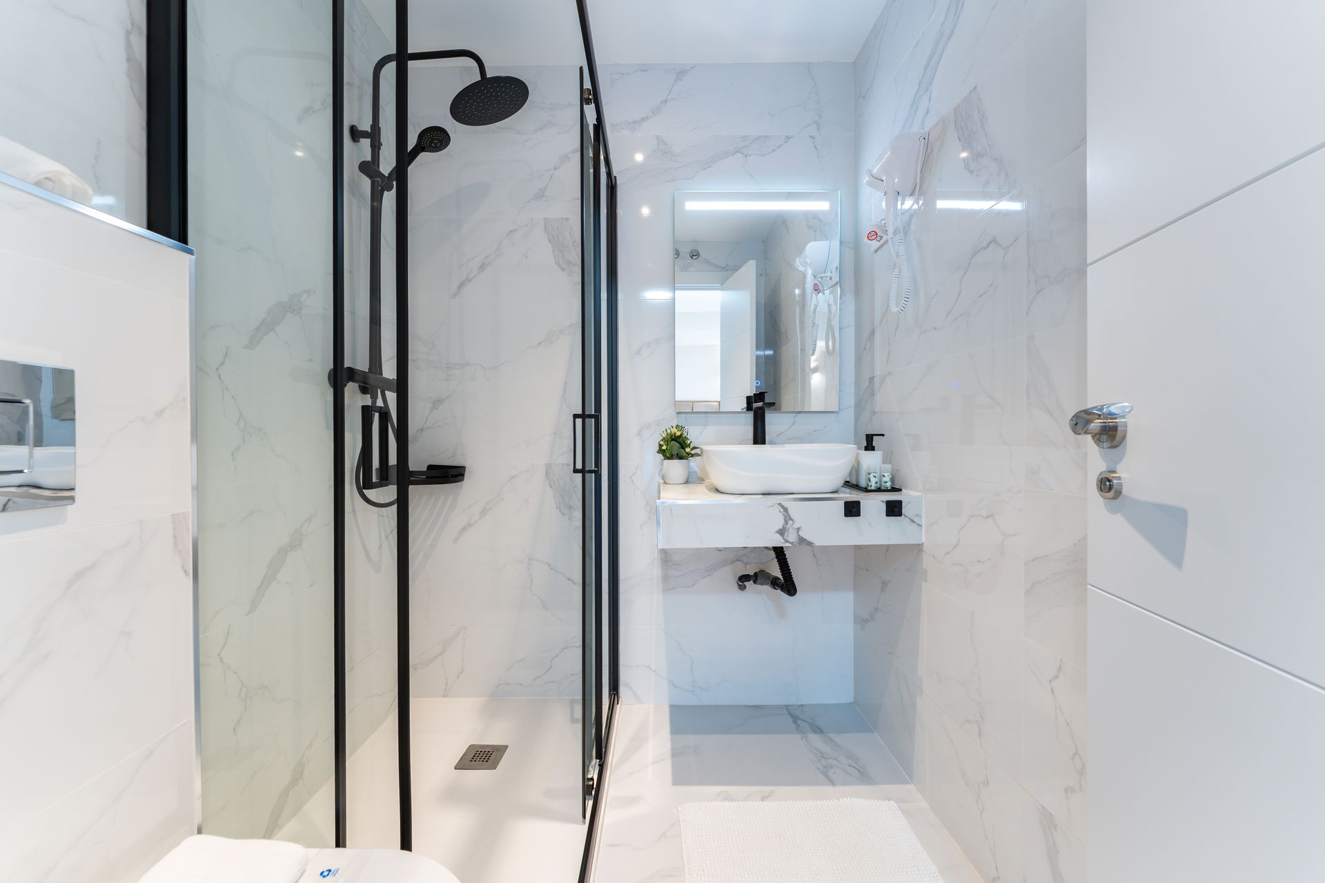 Baño moderno blanco con grifería de ducha negra, azulejos de mármol y lavabo suspendido.