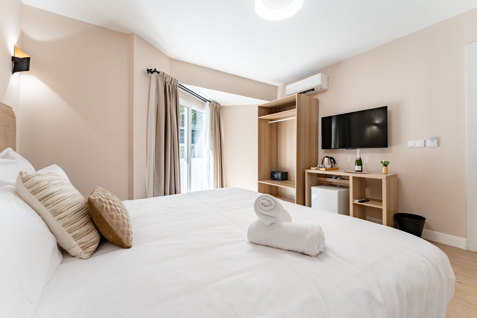 Dormitorio con cama grande, televisión, armario y nevera pequeña. Colores neutros, luz natural.