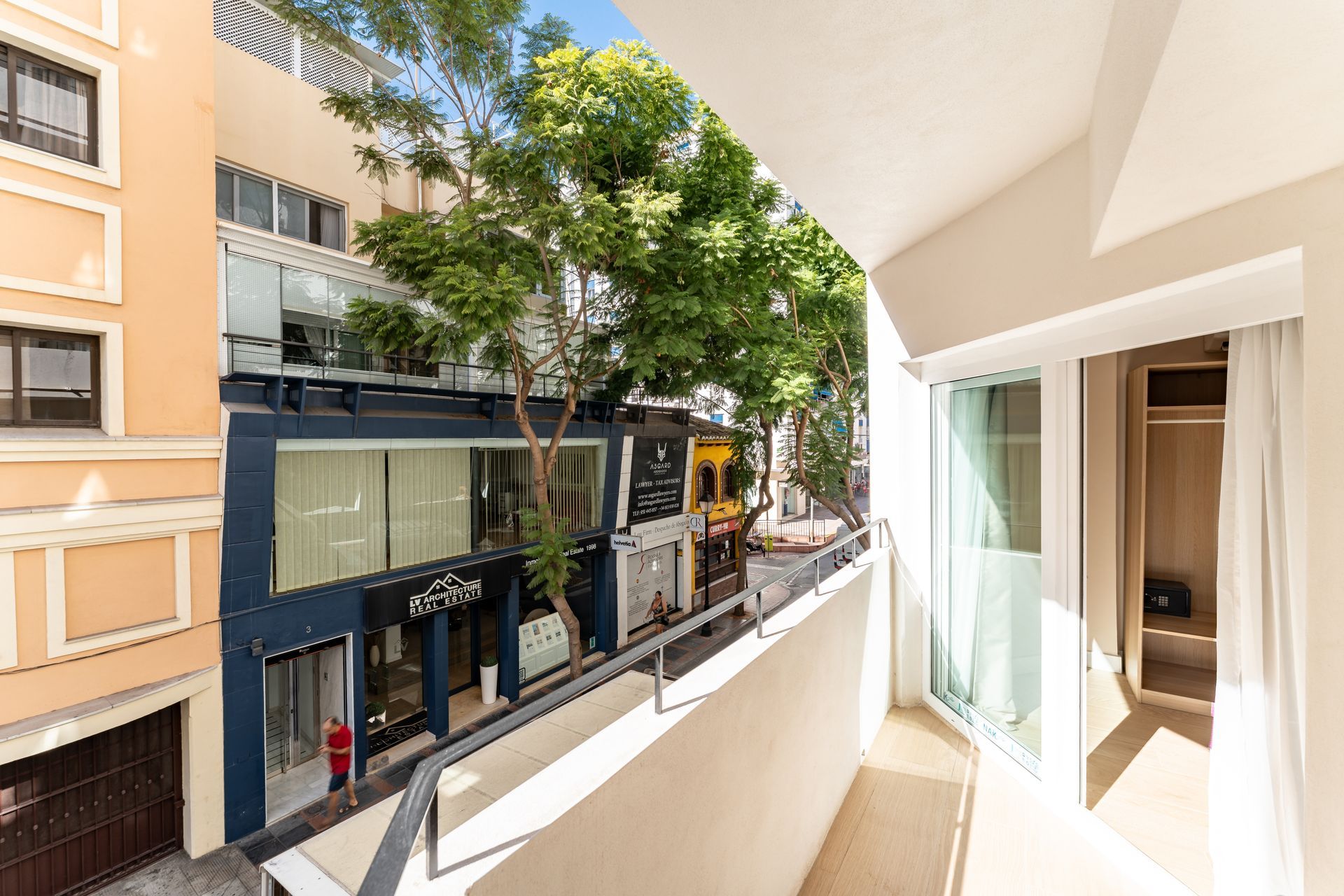 Vista desde el balcón a una calle con tiendas y un árbol; paredes de color beige claro, una puerta corredera de cristal