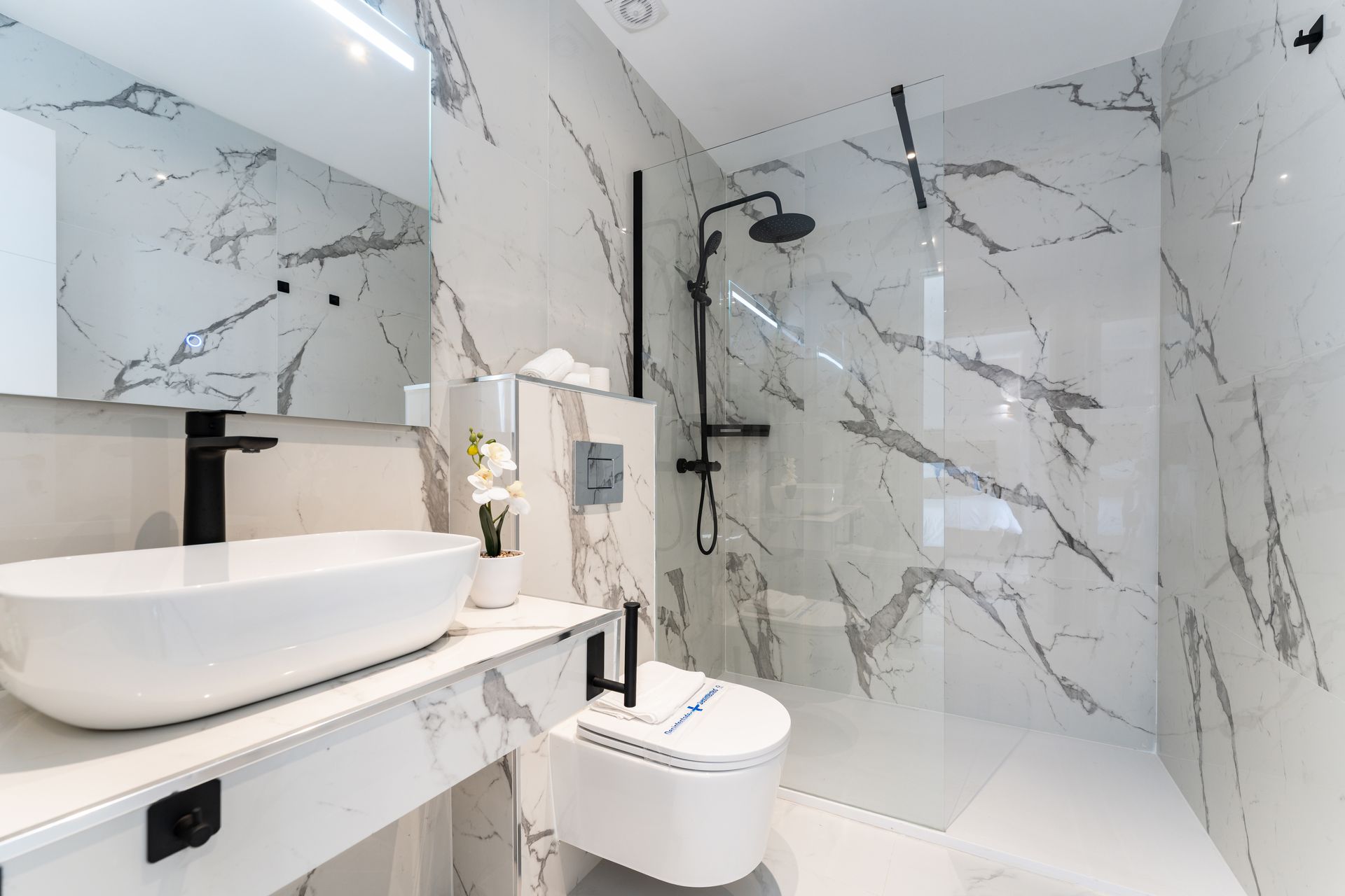 Baño moderno con paredes de mármol blanco, grifería negra y ducha de cristal.