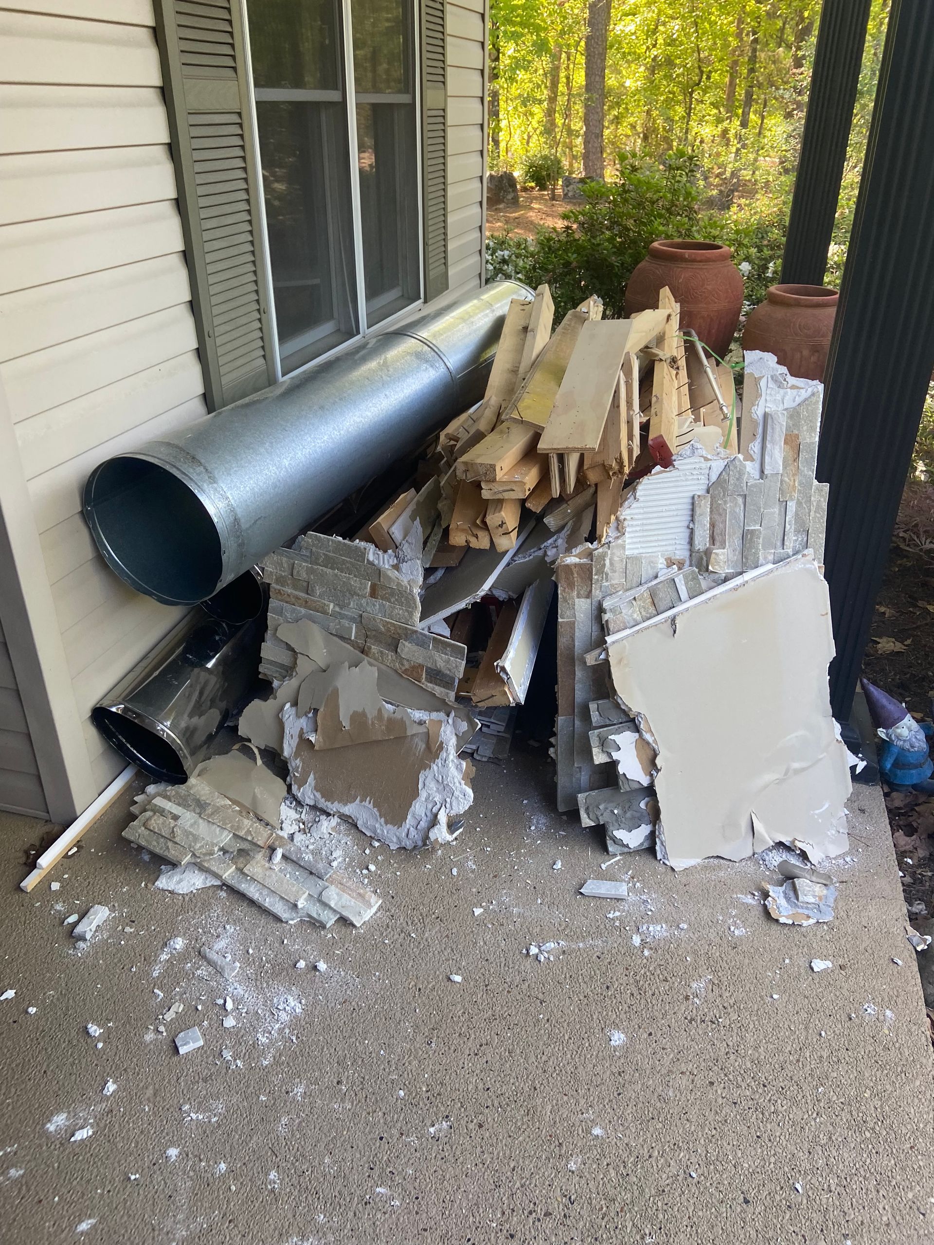 A person holding a long, beige board stands beside a pile of wood in an outdoor setting.