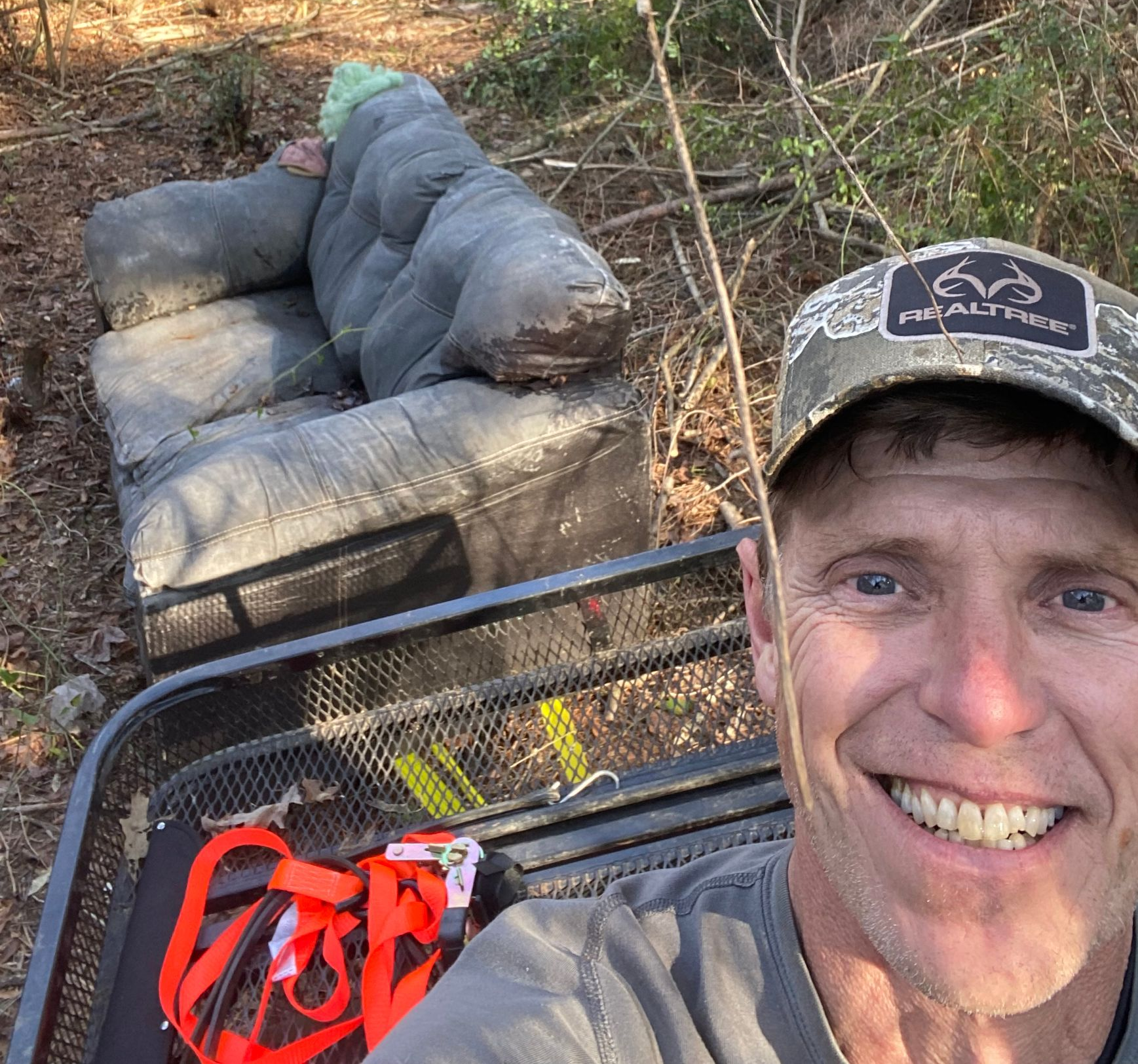 Man smiling, near a cart and discarded sofa in a wooded area.
