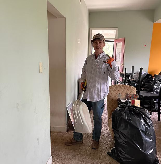 Man holding a shovel and a trash bag, standing in a doorway. Room has debris and is being cleaned.