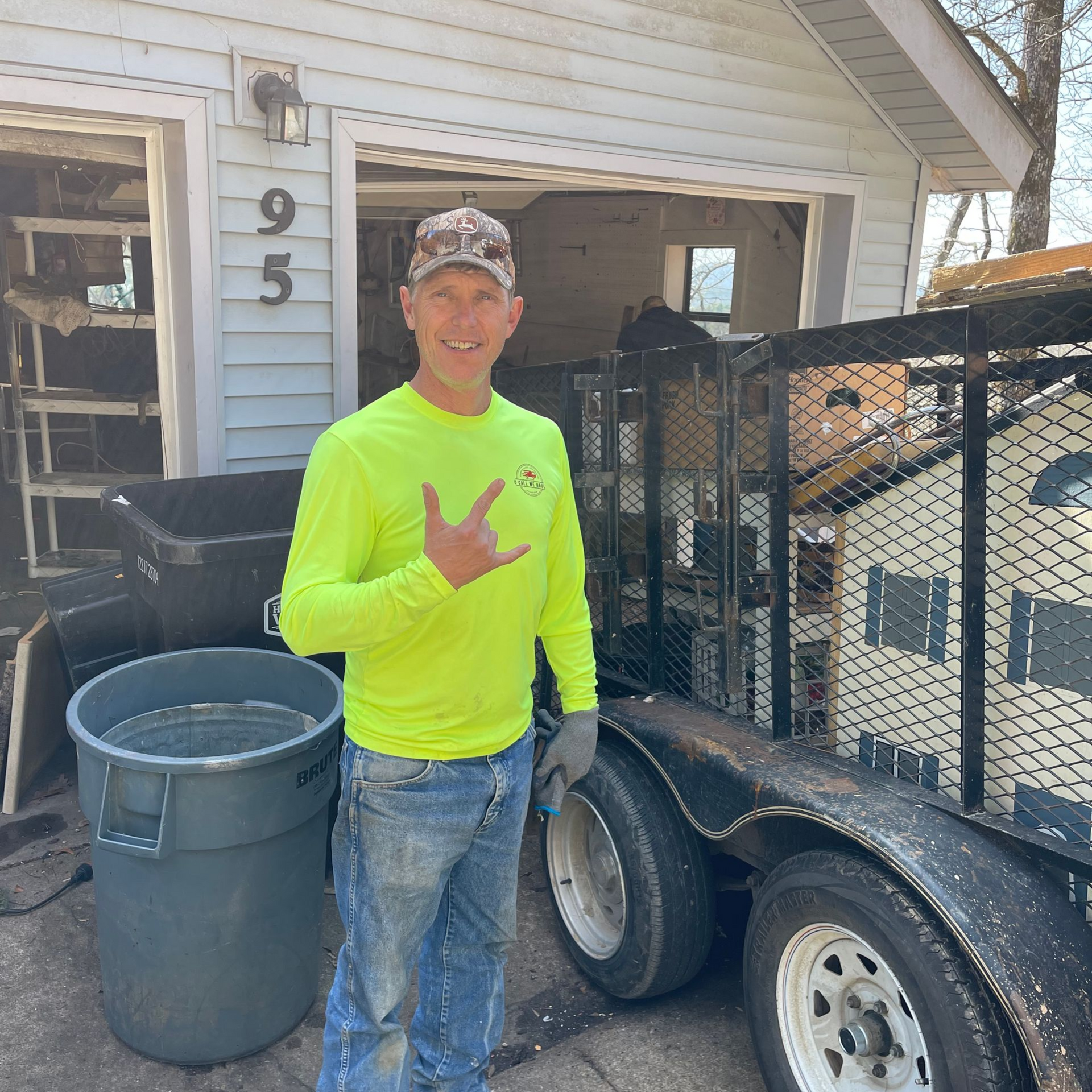 Man in neon shirt and hat giving a hand gesture, standing beside a trailer filled with items in front of a house.