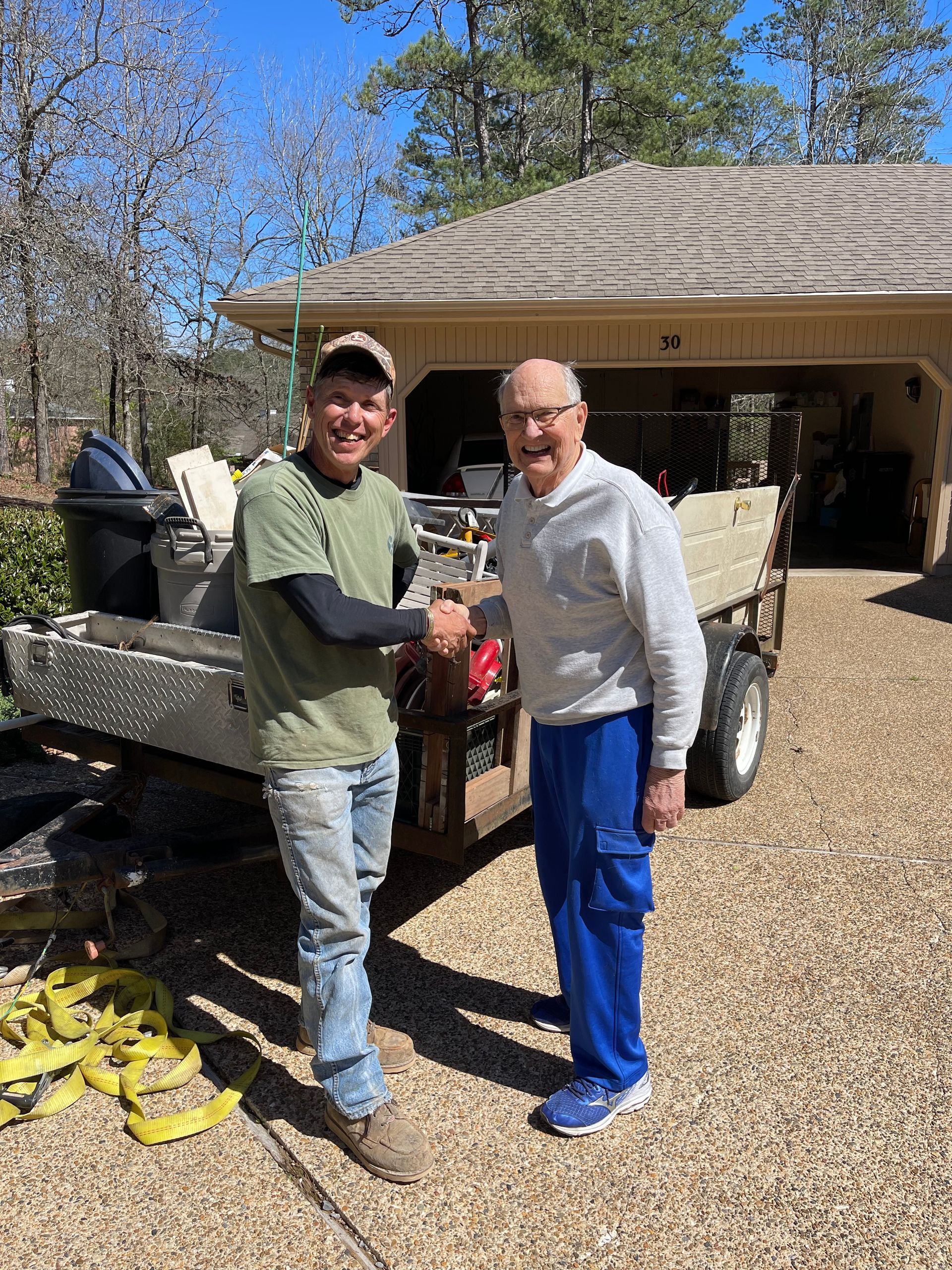 Man in green shirt shakes hands with an older man in blue pants near a trailer, sunny day.