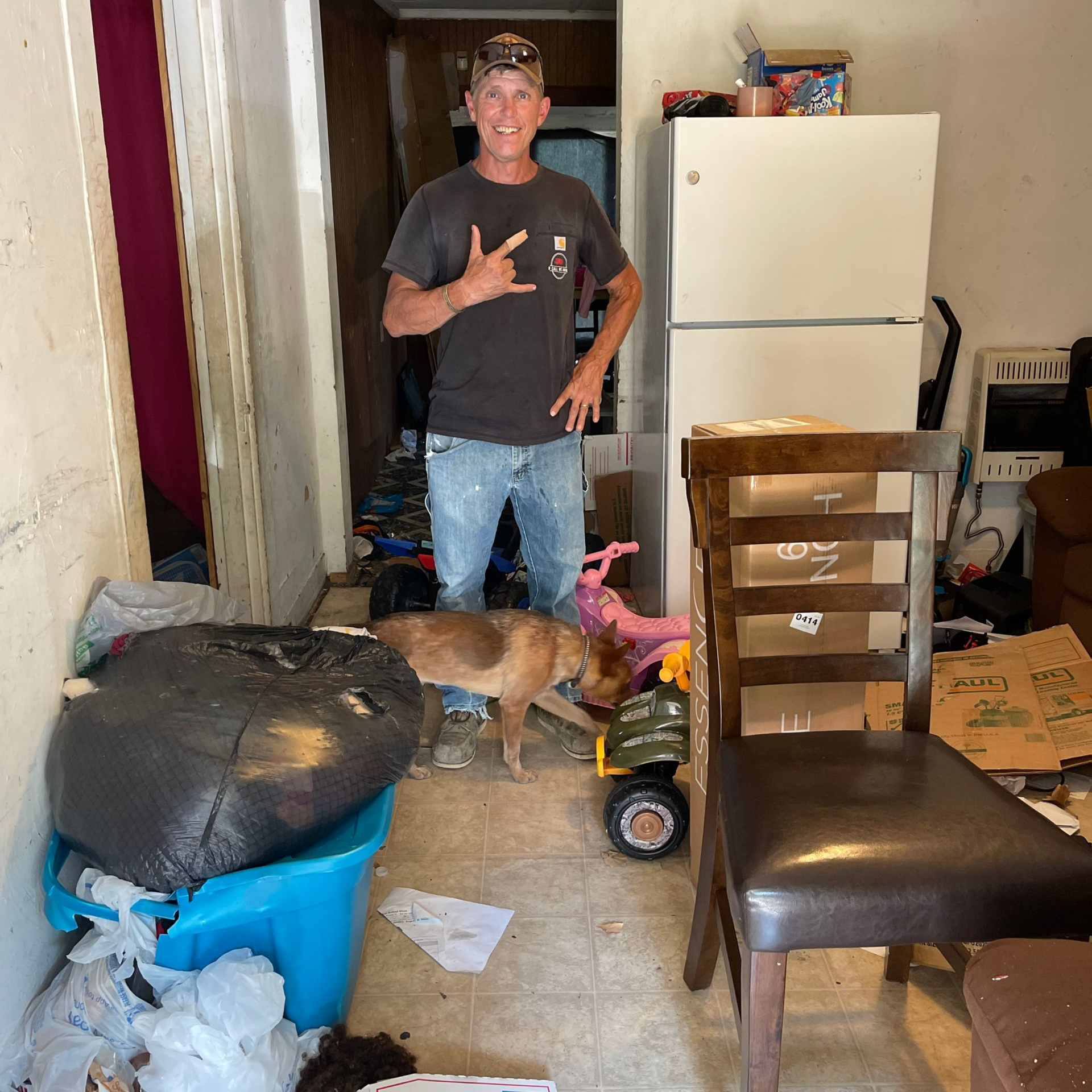 Man in messy room gestures, dog nearby. Trash, fridge, chair visible.