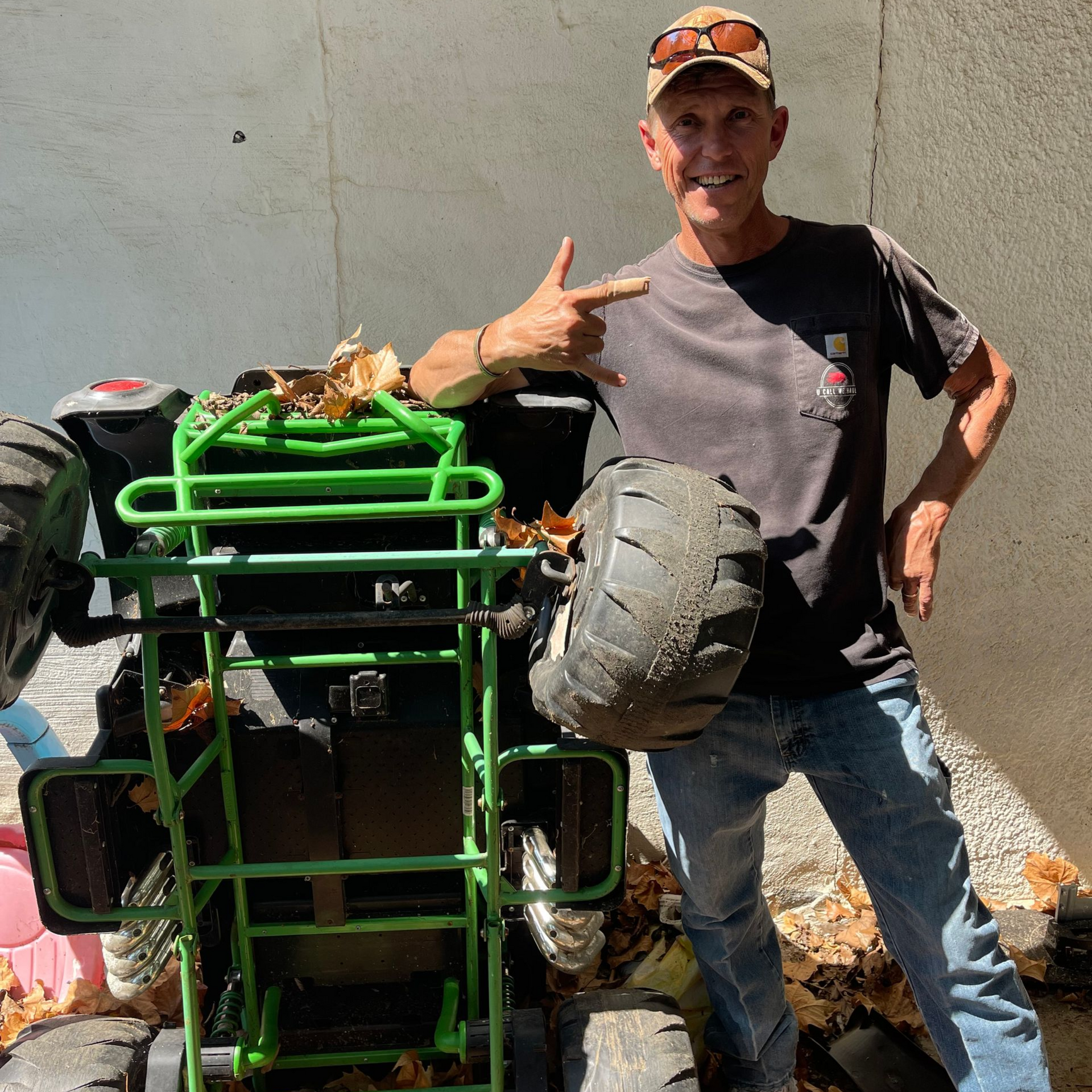 Man smiles, points at small green go-kart. Outdoor setting, leaves on ground.
