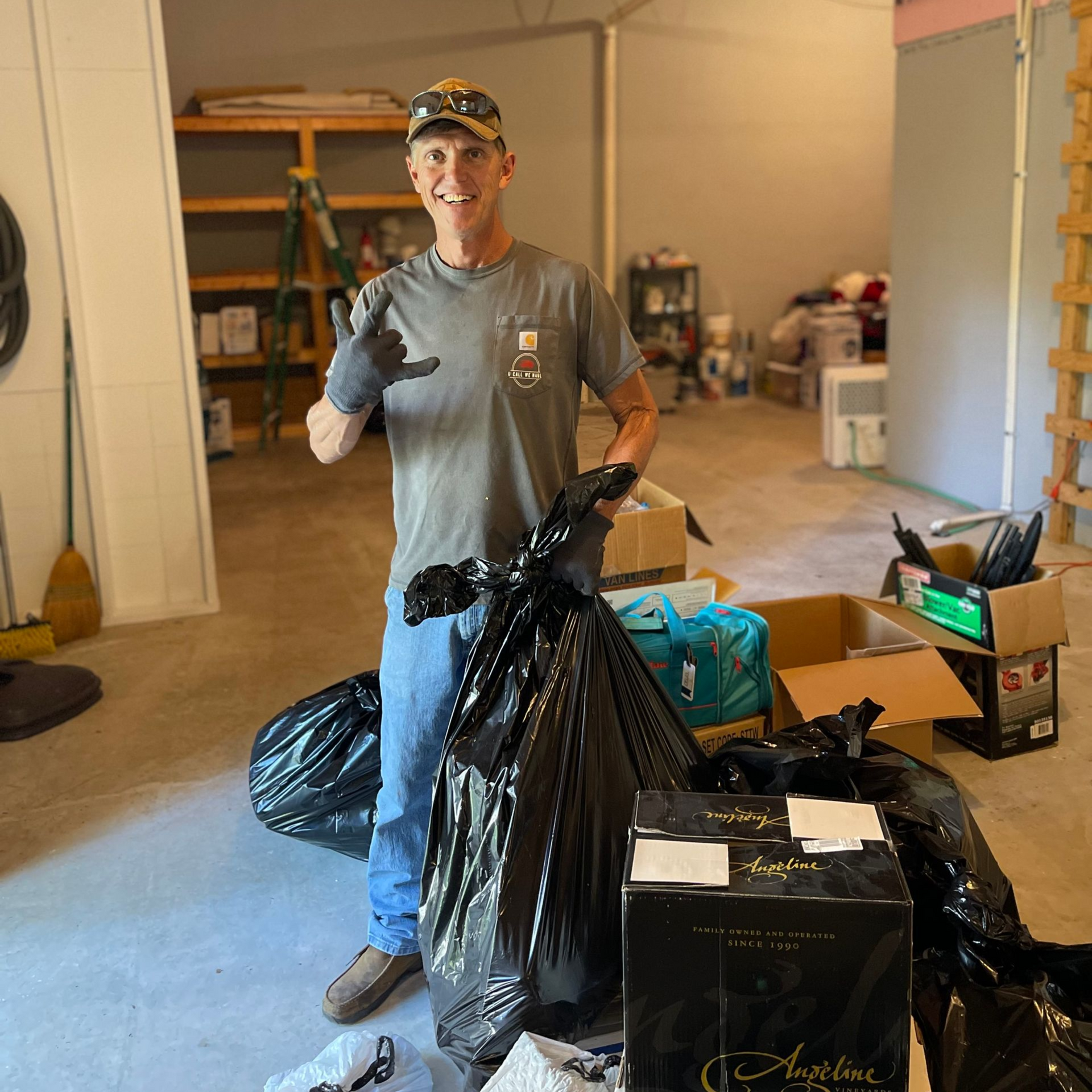 Man in cap and gloves holding trash bags in a garage, giving a shaka sign.