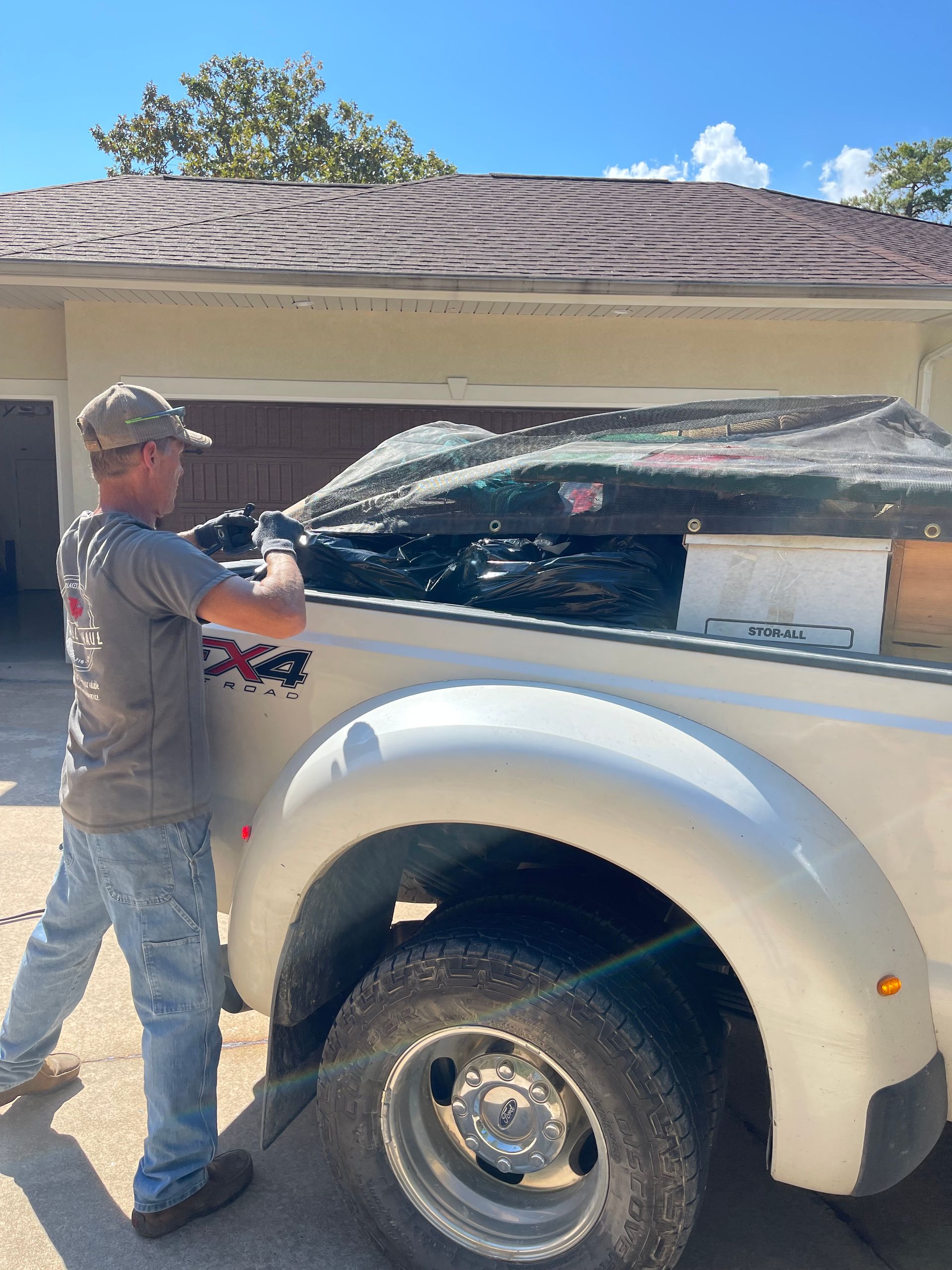 Man secures tarp over truck bed full of items, next to a building with brown roof, sunny day.
