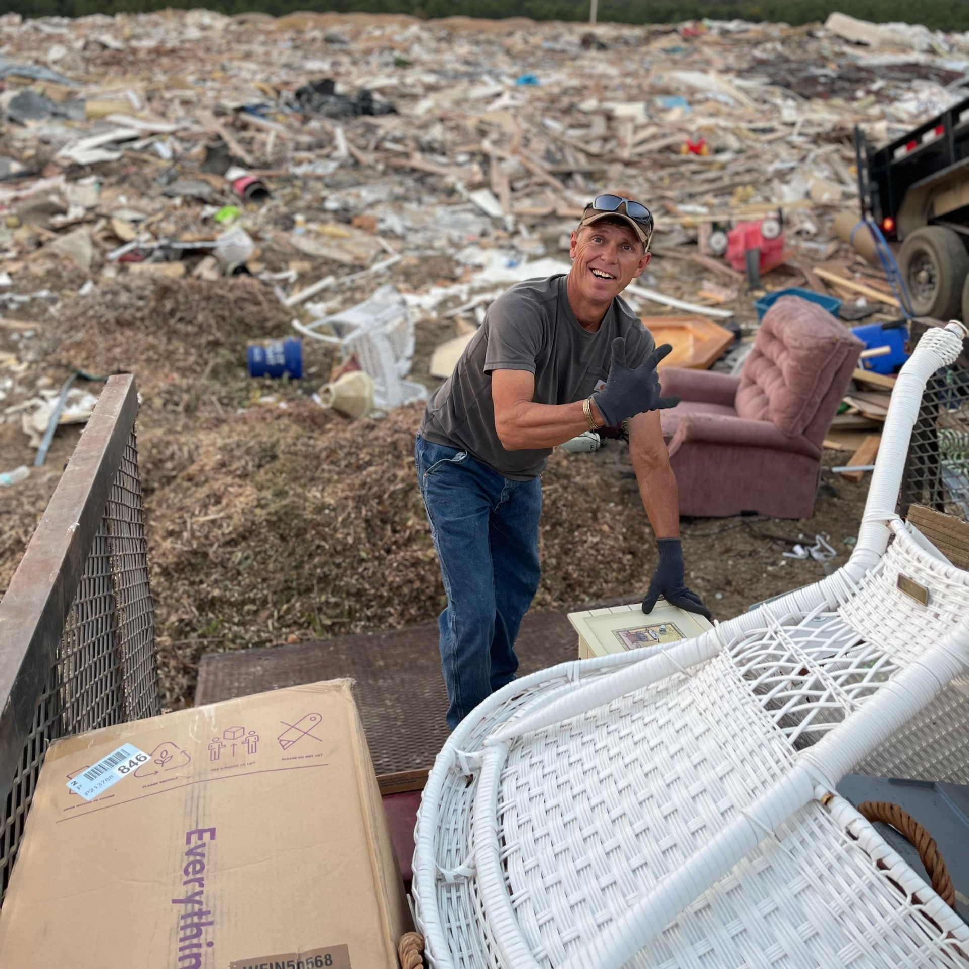 Man smiles, unloading items from a truck at a debris site. Items include furniture and a large cardboard box.