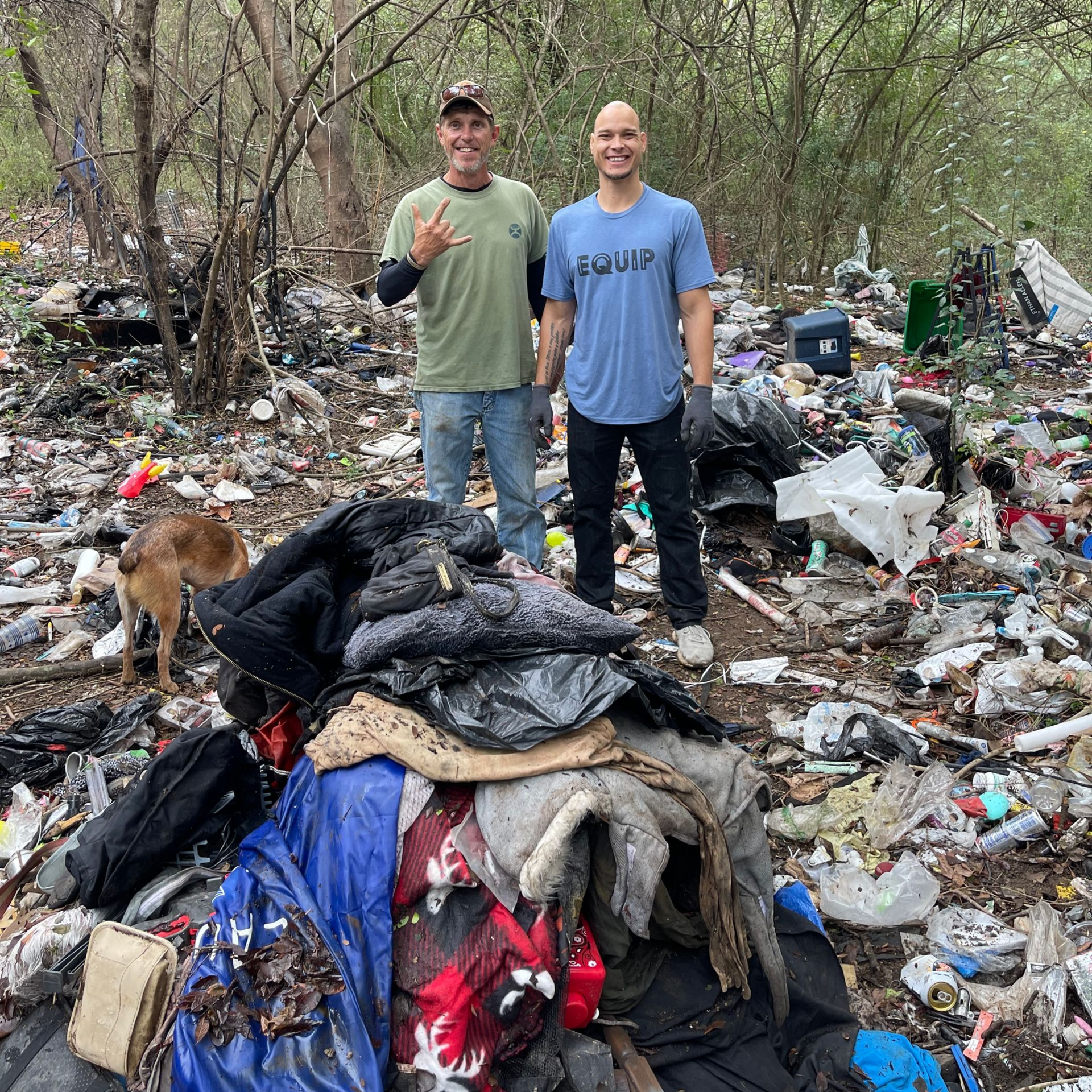 Two men stand smiling amidst a large pile of trash in a wooded area.