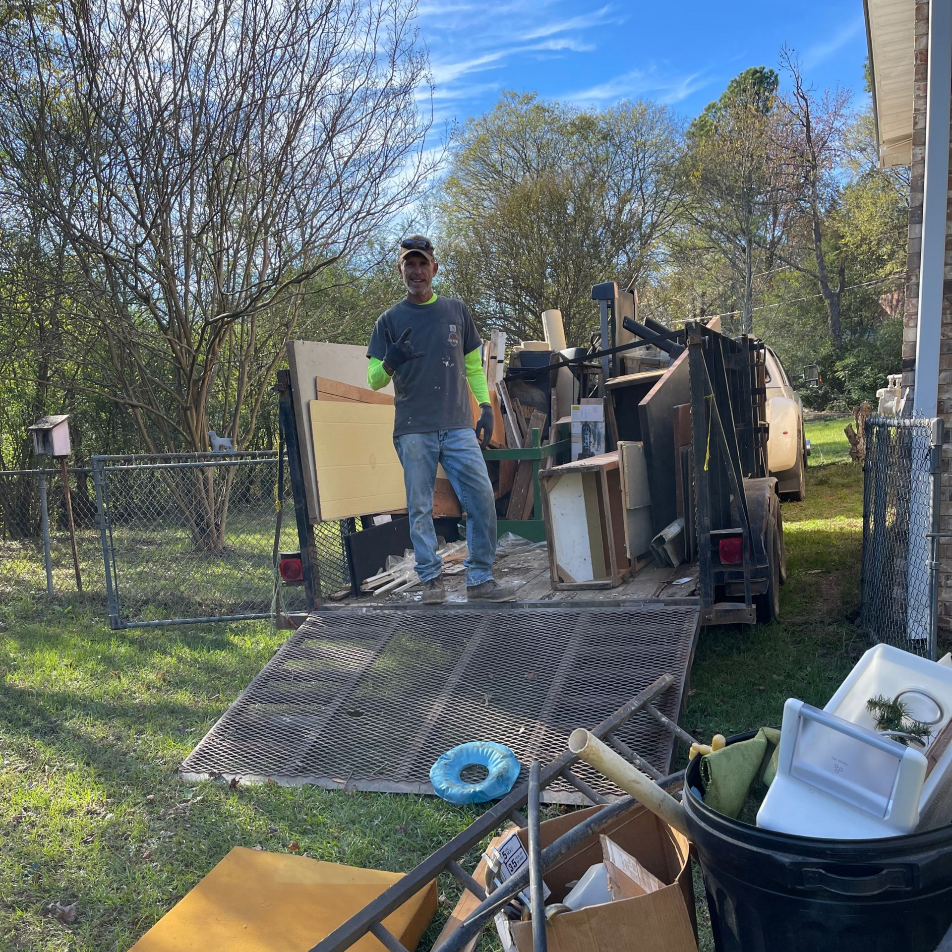 A man in a yard stands on a trailer full of debris. He is giving a thumbs up.