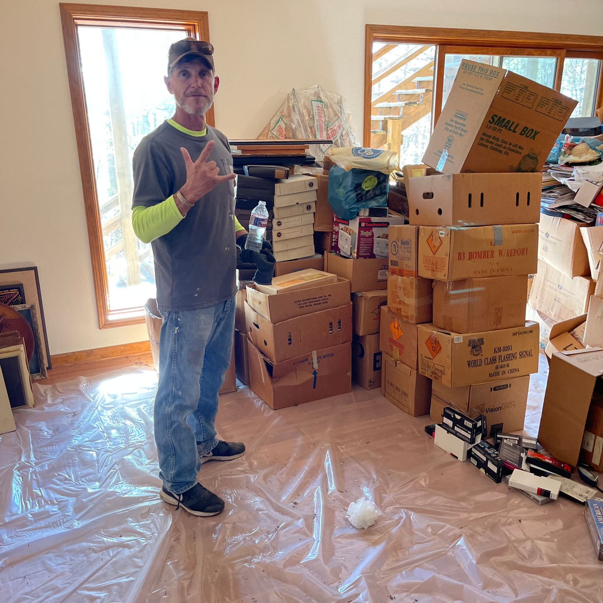 Man in casual clothes giving a peace sign, standing in a room surrounded by moving boxes.
