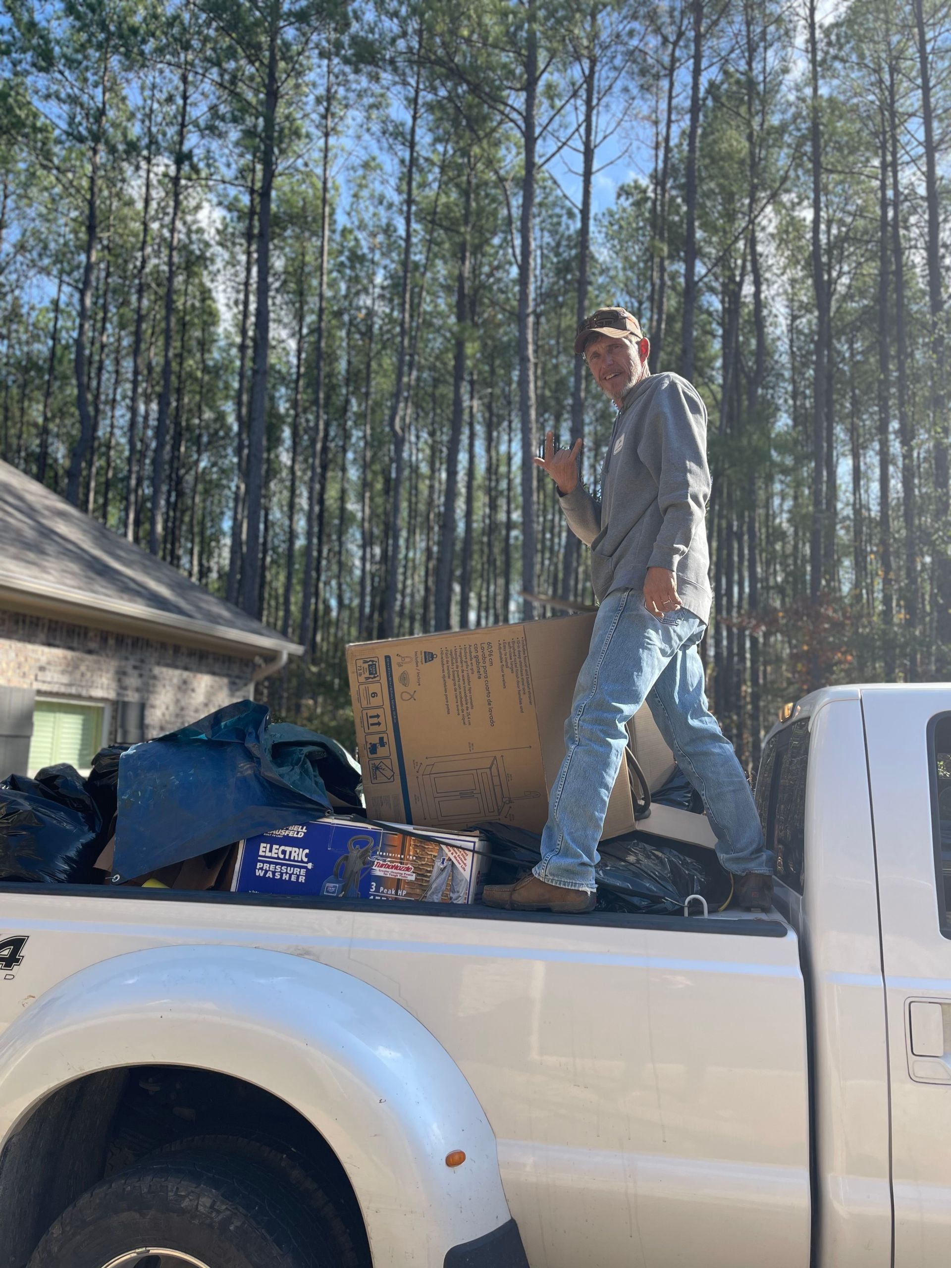 Man standing on truck bed, giving thumbs up. Truck filled with boxes and items, sunny day in front of trees.