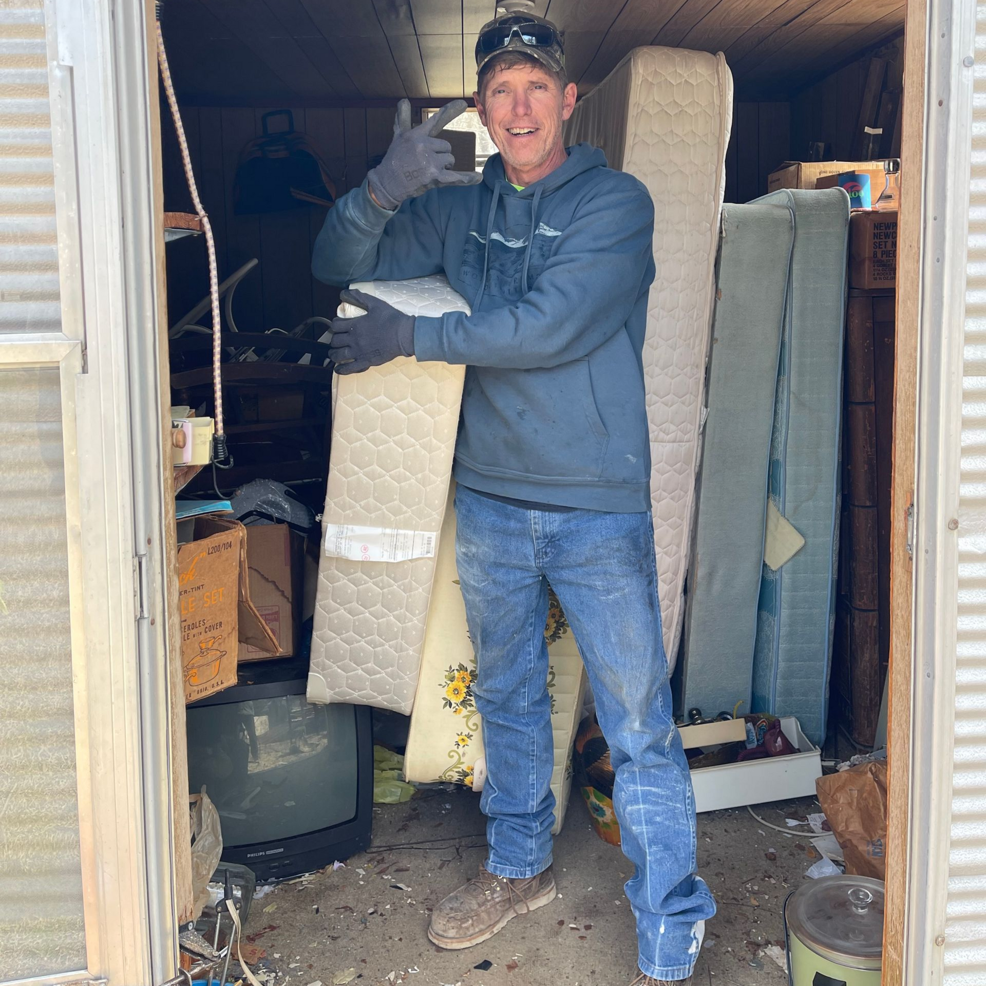 Man in blue hoodie and jeans gives thumbs up, holding mattress pieces in cluttered shed.