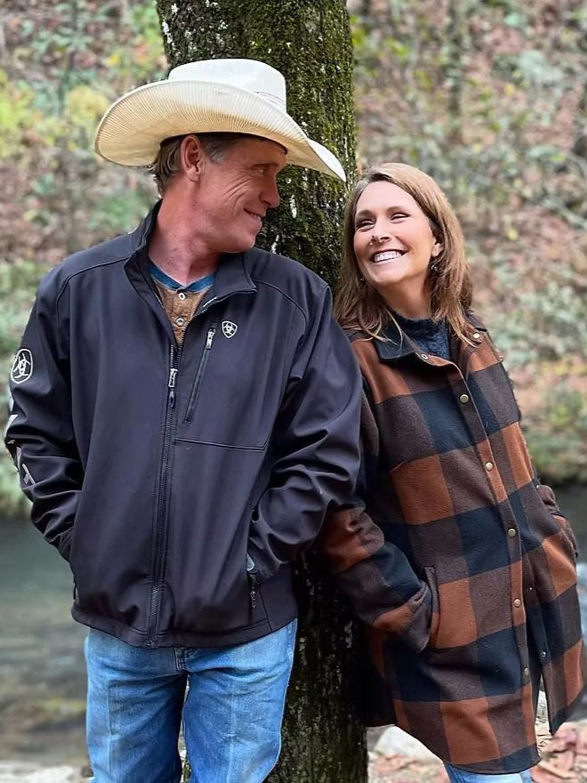 A couple in outdoor setting, smiling at each other. Man in cowboy hat, woman in plaid jacket, leaning against a tree.