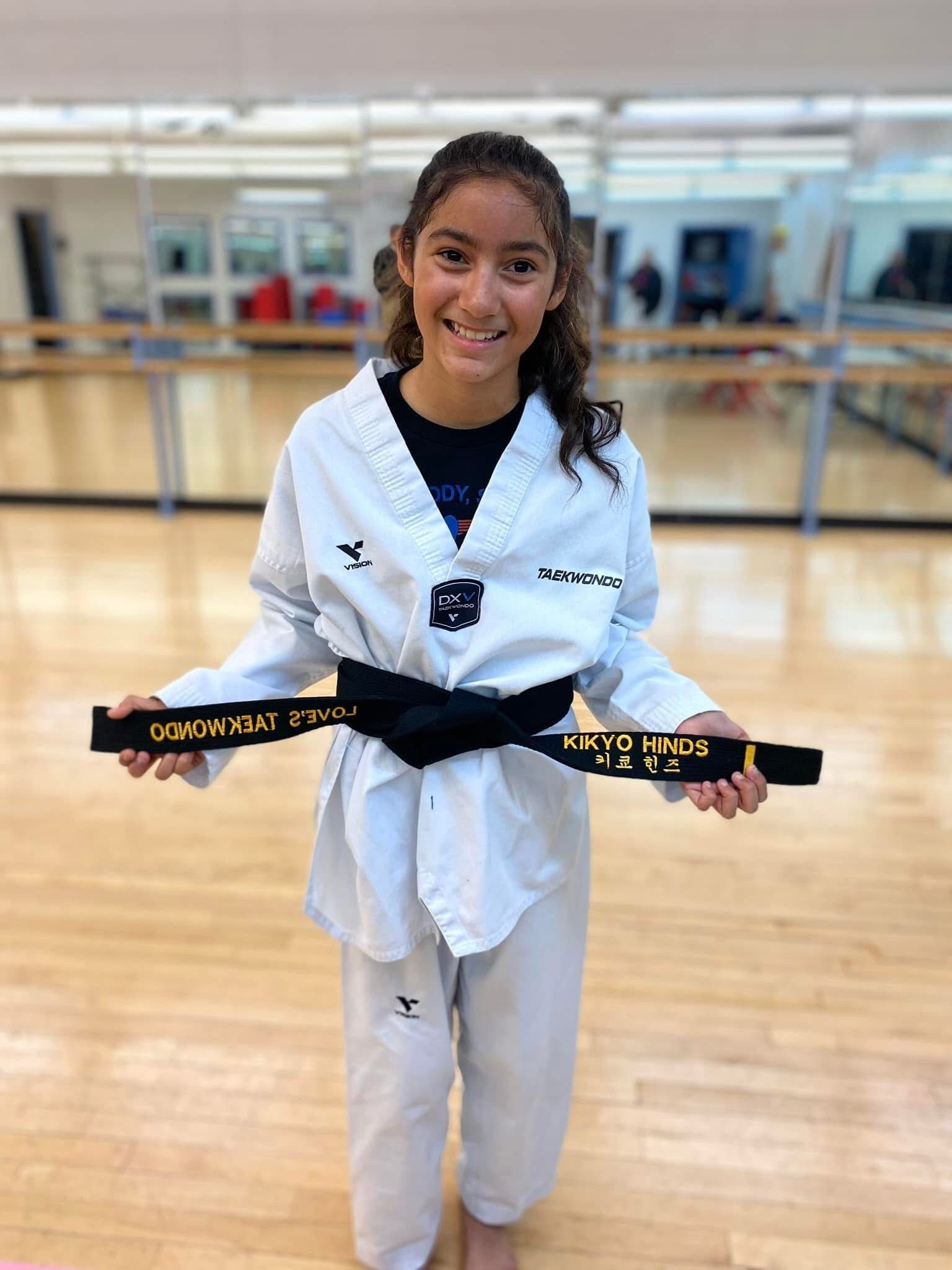 A young girl in a taekwondo uniform is holding a black belt in a gym.
