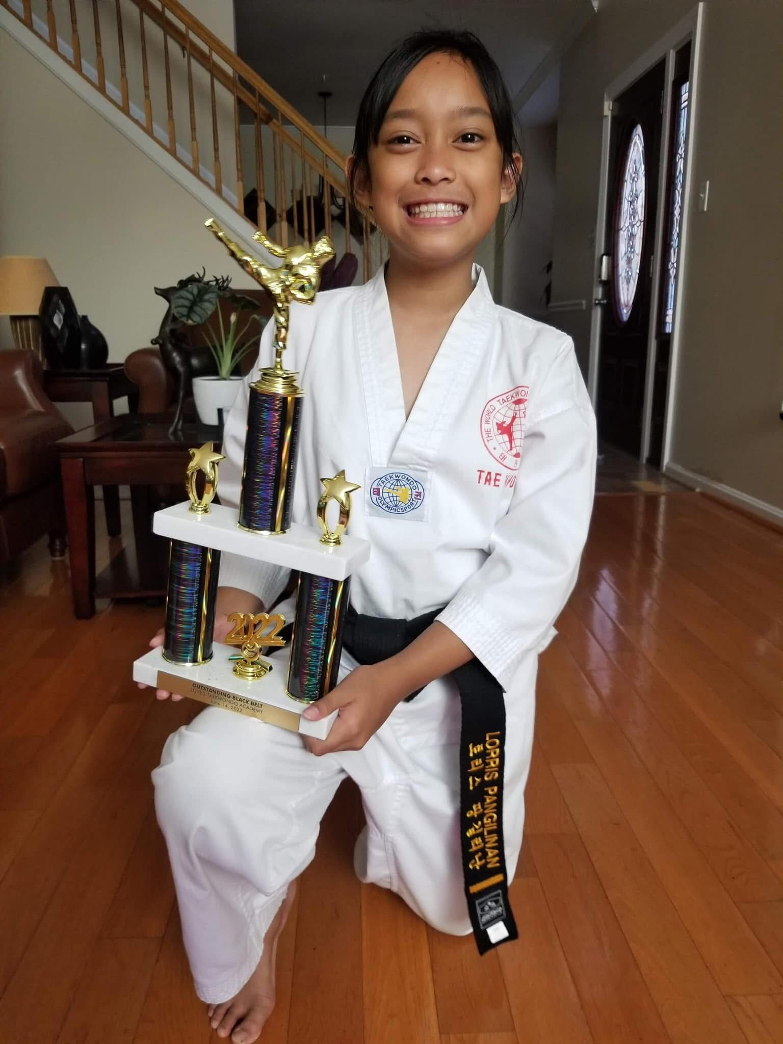 A young girl in a karate uniform is kneeling down holding a trophy.