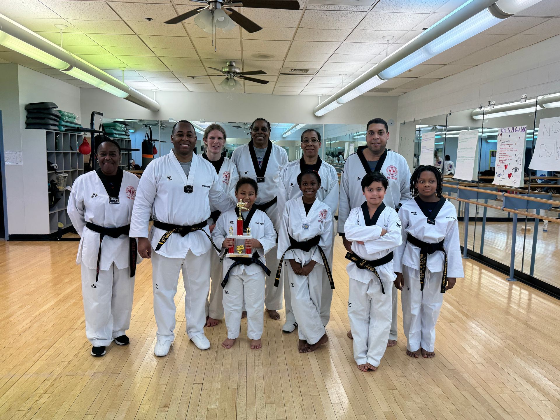 Group of Taekwondo practitioners in white uniforms, posing with a trophy in a dojo.