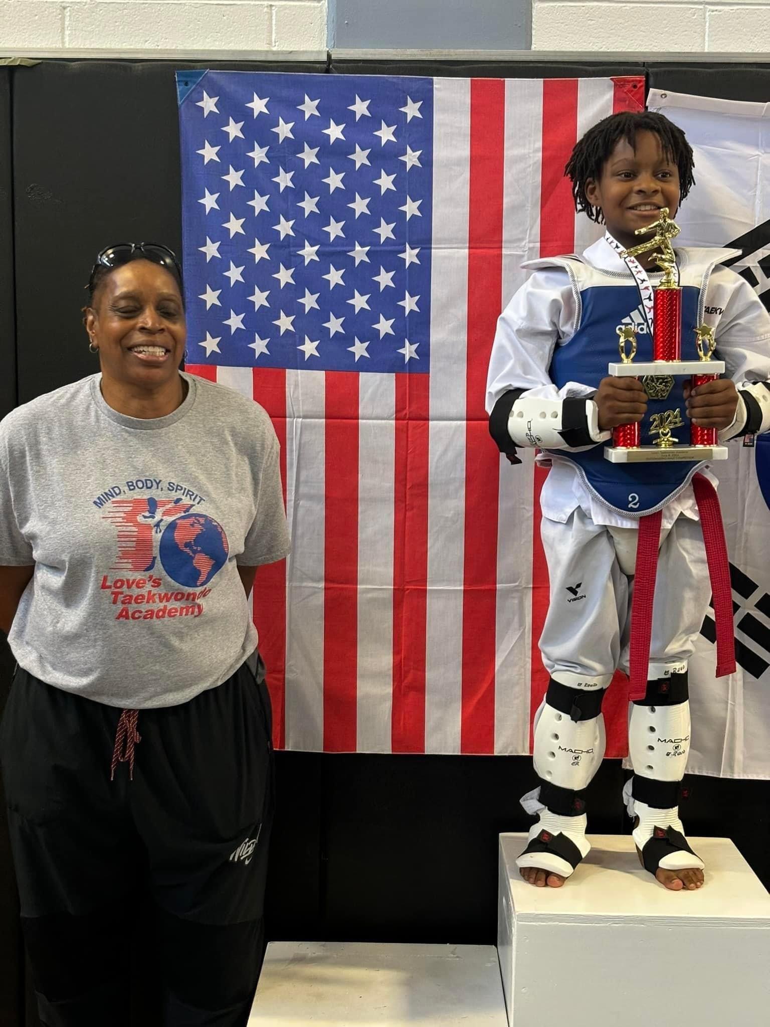 A woman is standing next to a boy holding a trophy in front of an american flag.