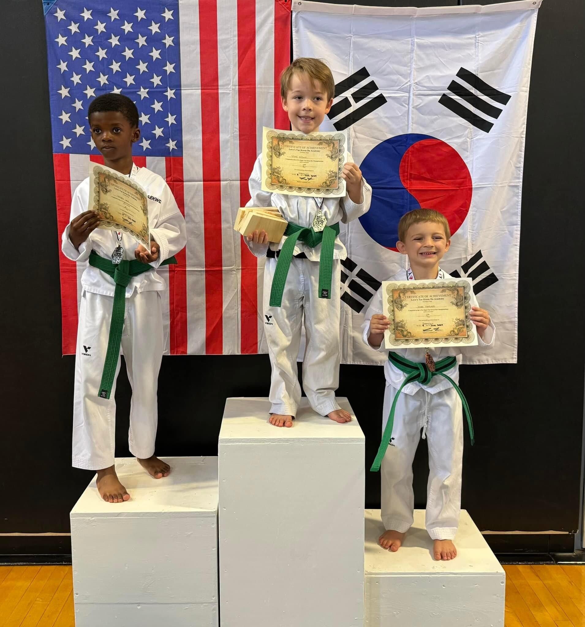 Three young boys are standing on a podium holding certificates in front of a flag.