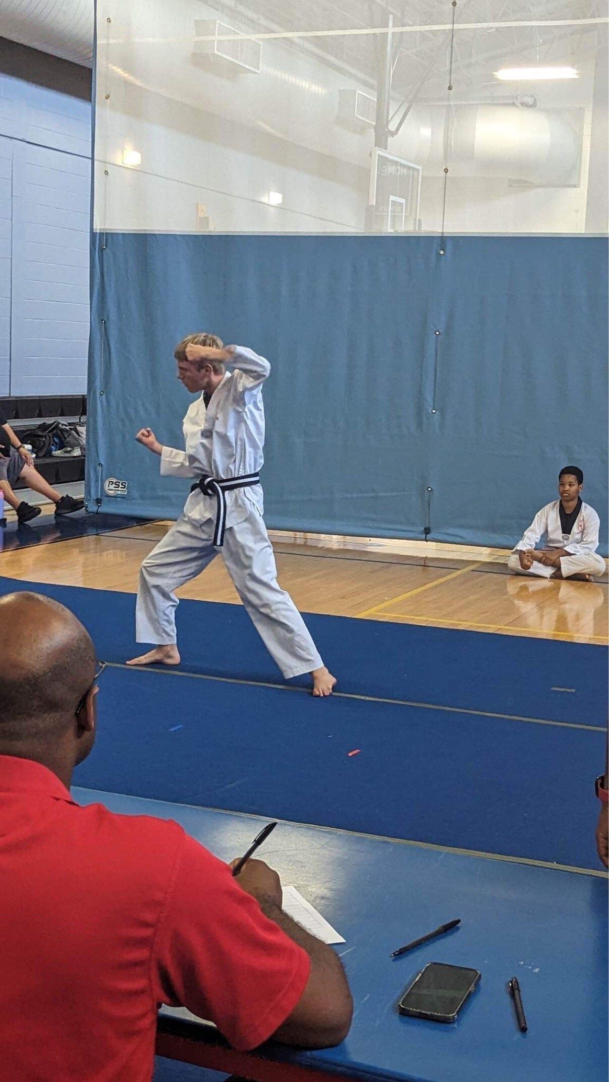 A man in a red shirt is sitting at a table in a gym watching a karate match.
