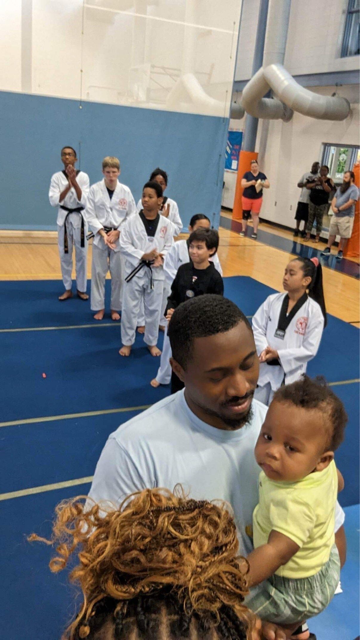A man is holding a baby in front of a group of people standing in a gym.