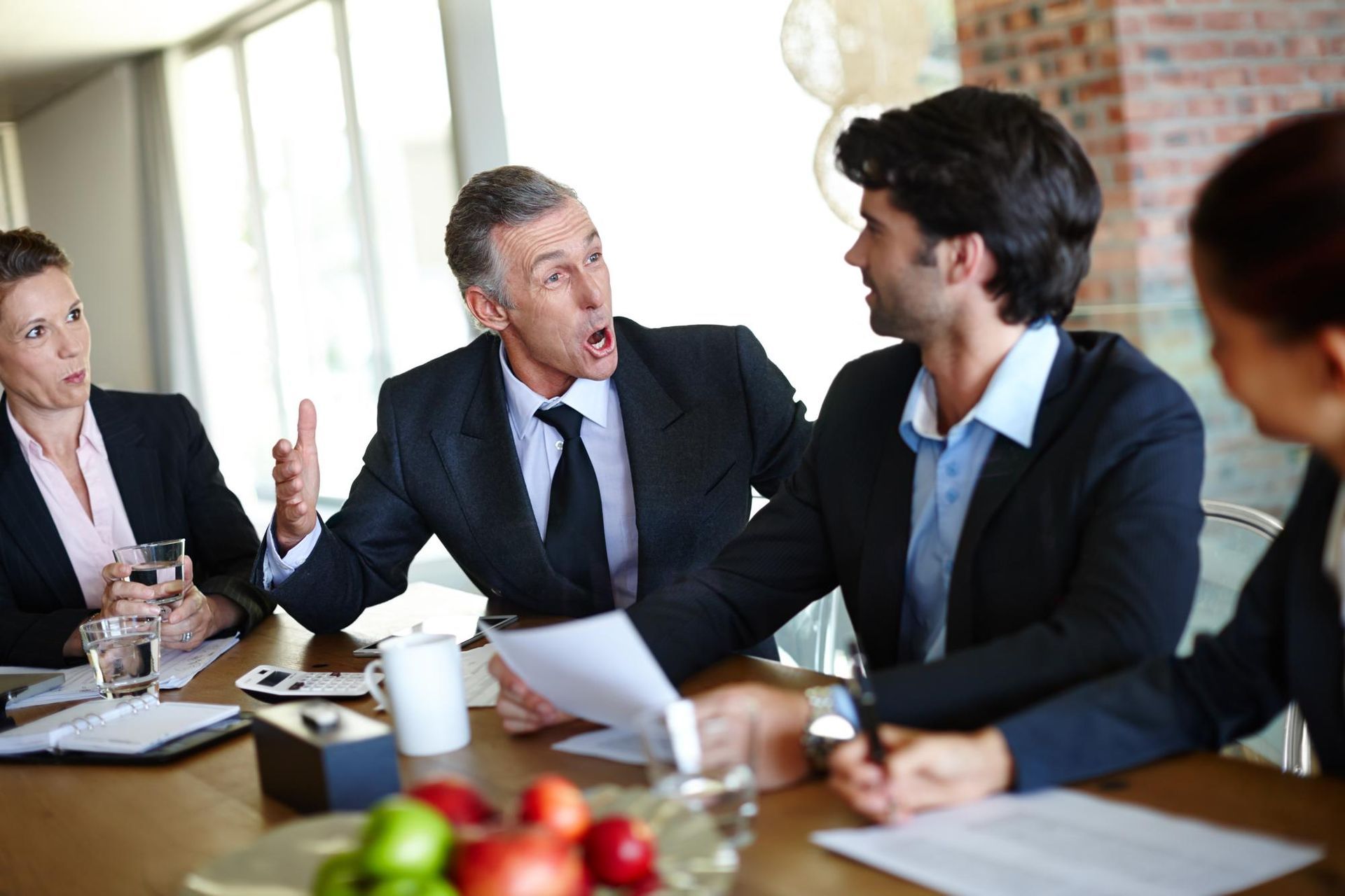 Business meeting. Man gestures angrily, papers in hand. Others look on, some concerned.