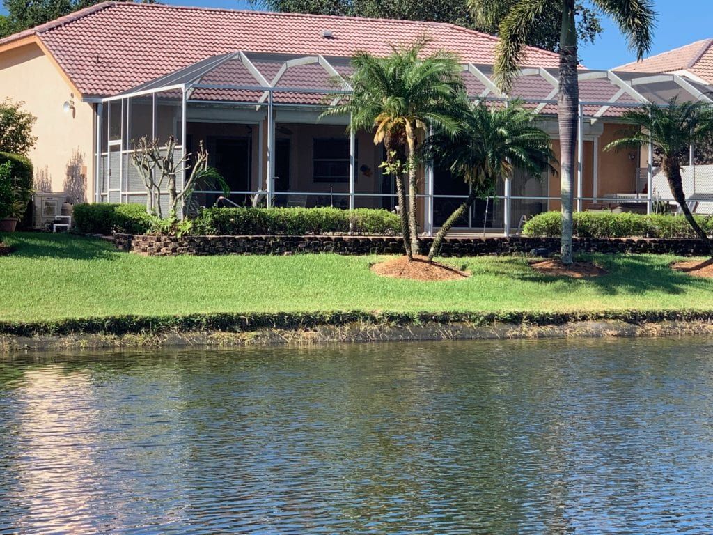 House with screened porch by a lake, green grass, palm trees.