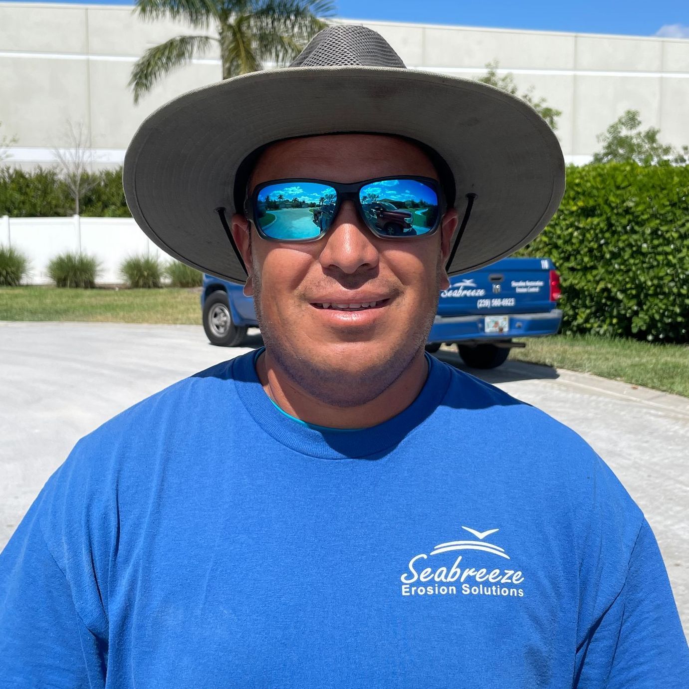 Man in blue shirt, sunglasses, and hat smiles outdoors, a blue van behind him.