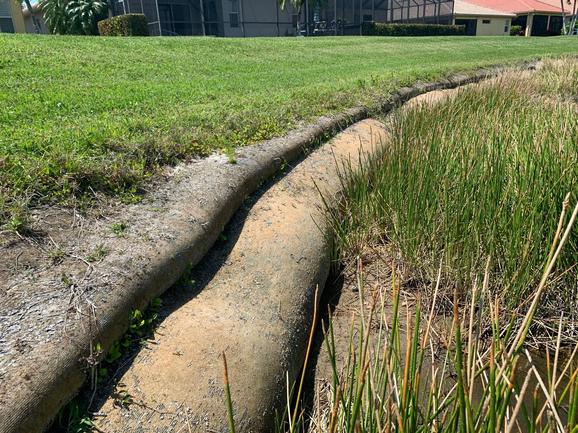 A dry drainage ditch with a sandbag-like barrier between green grass and reeds, in a residential yard.