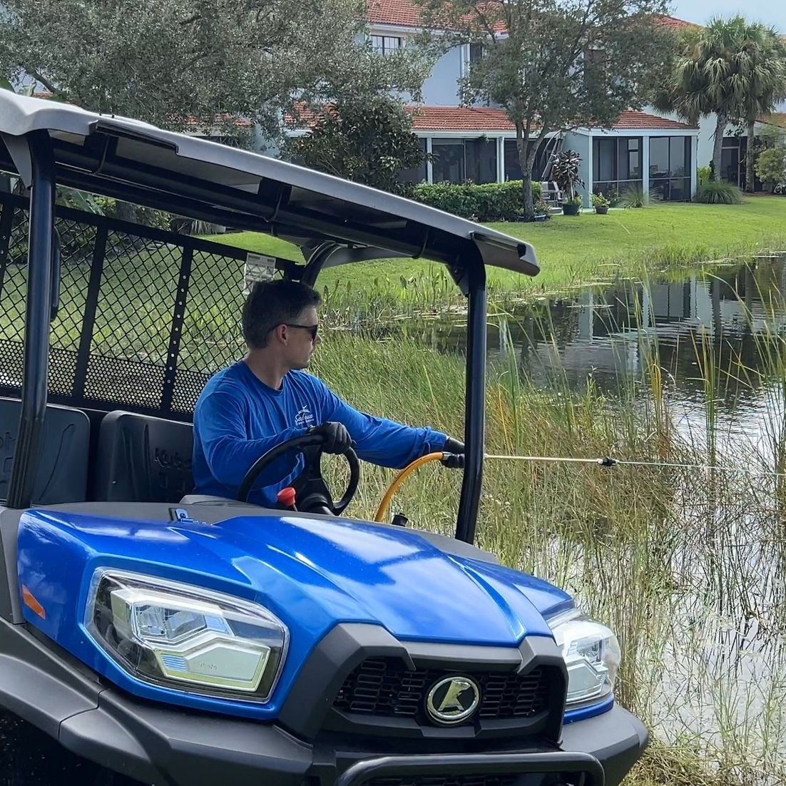 Man in blue shirt driving blue utility vehicle, spraying weeds near a lake.