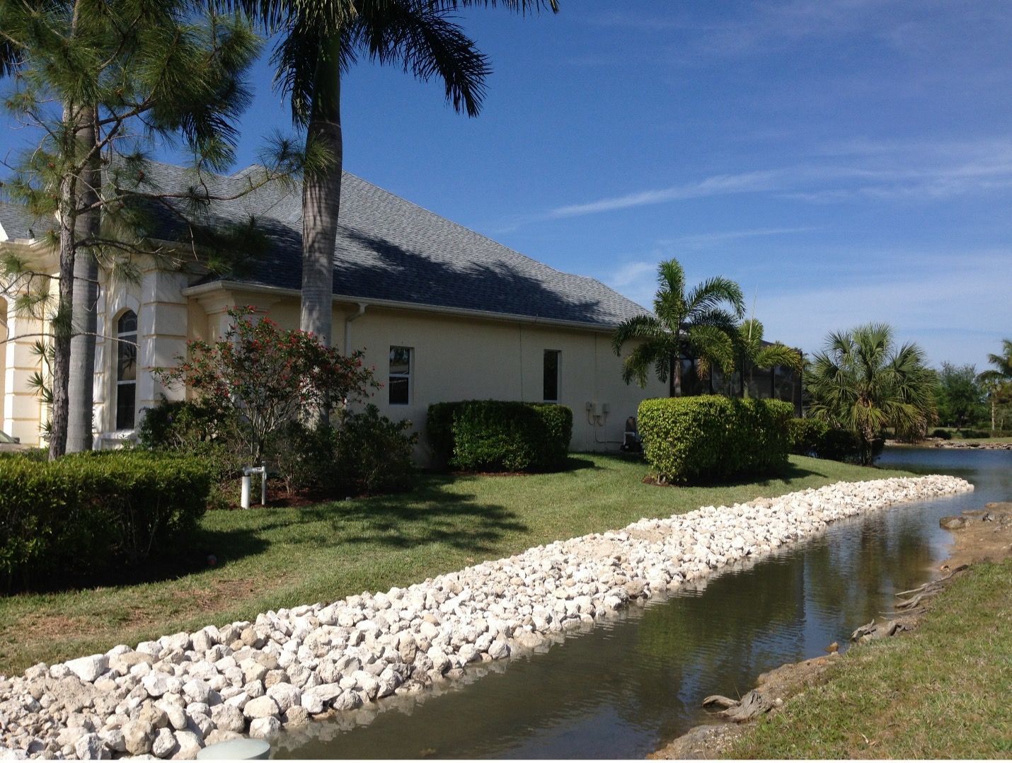 House beside a waterway, with rock bank and lawn, under a blue sky.